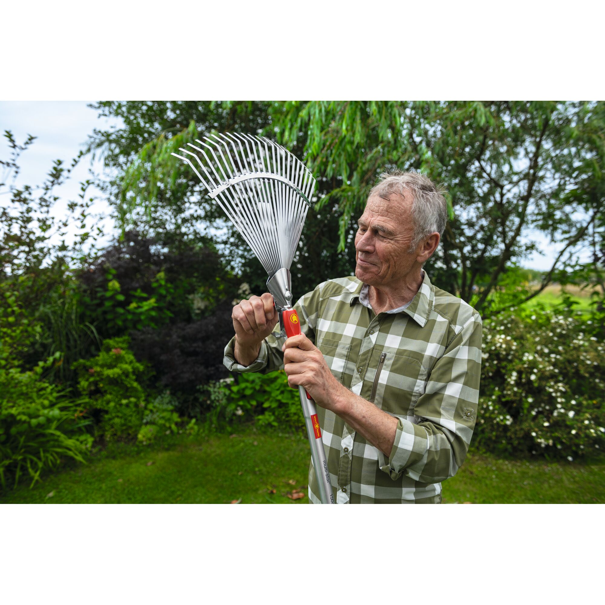A person outdoors in a garden holding and adjusting a WOLFGarten UE-M adjustable rake with a metal head. The person's face is blurred for privacy.