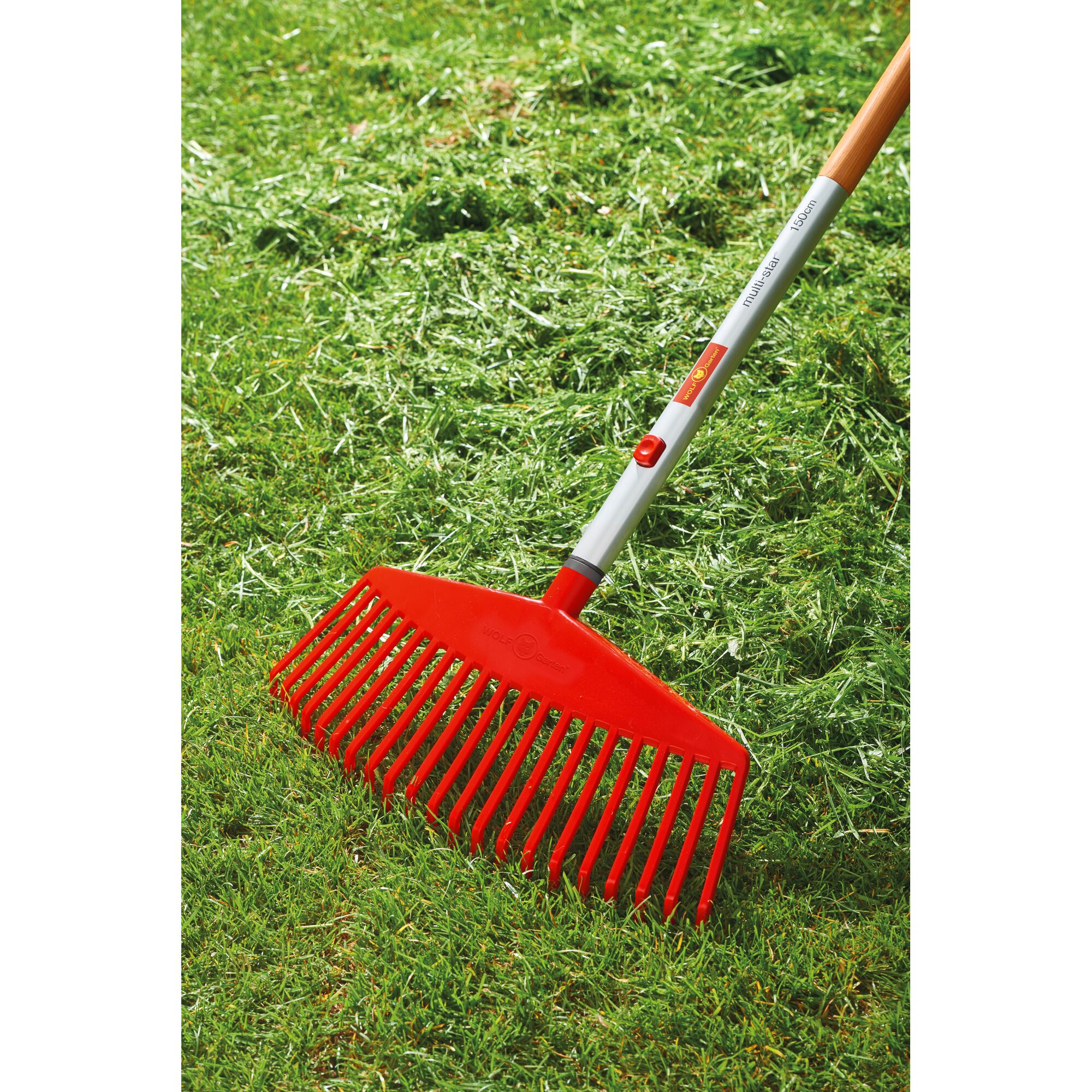 A red WOLF-Garten lawn rake with a wooden handle is shown being used to gather grass clippings on a green lawn.