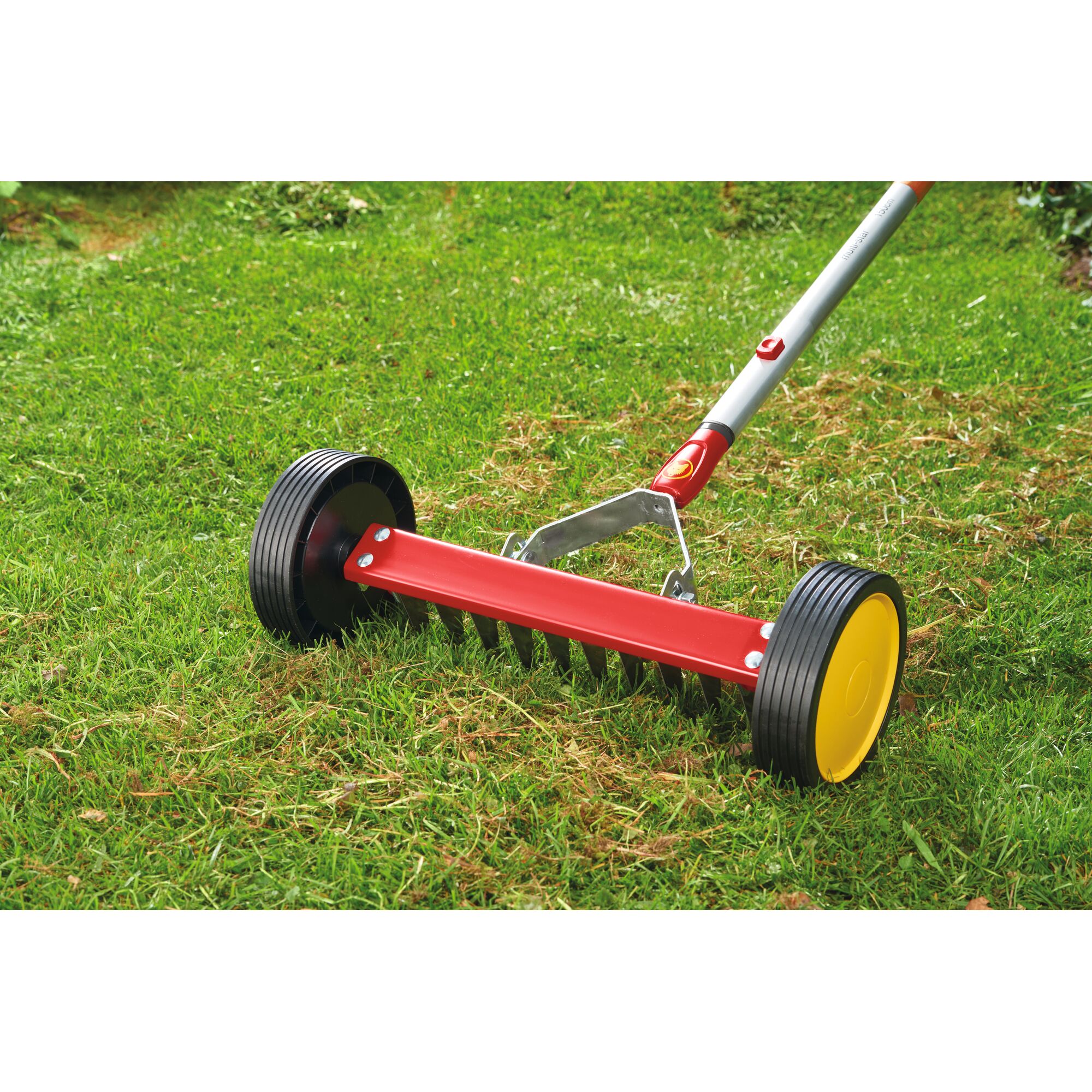 A close-up of a lawn scarifier with a red body, black wheels, and yellow wheel centers being used on grass.
