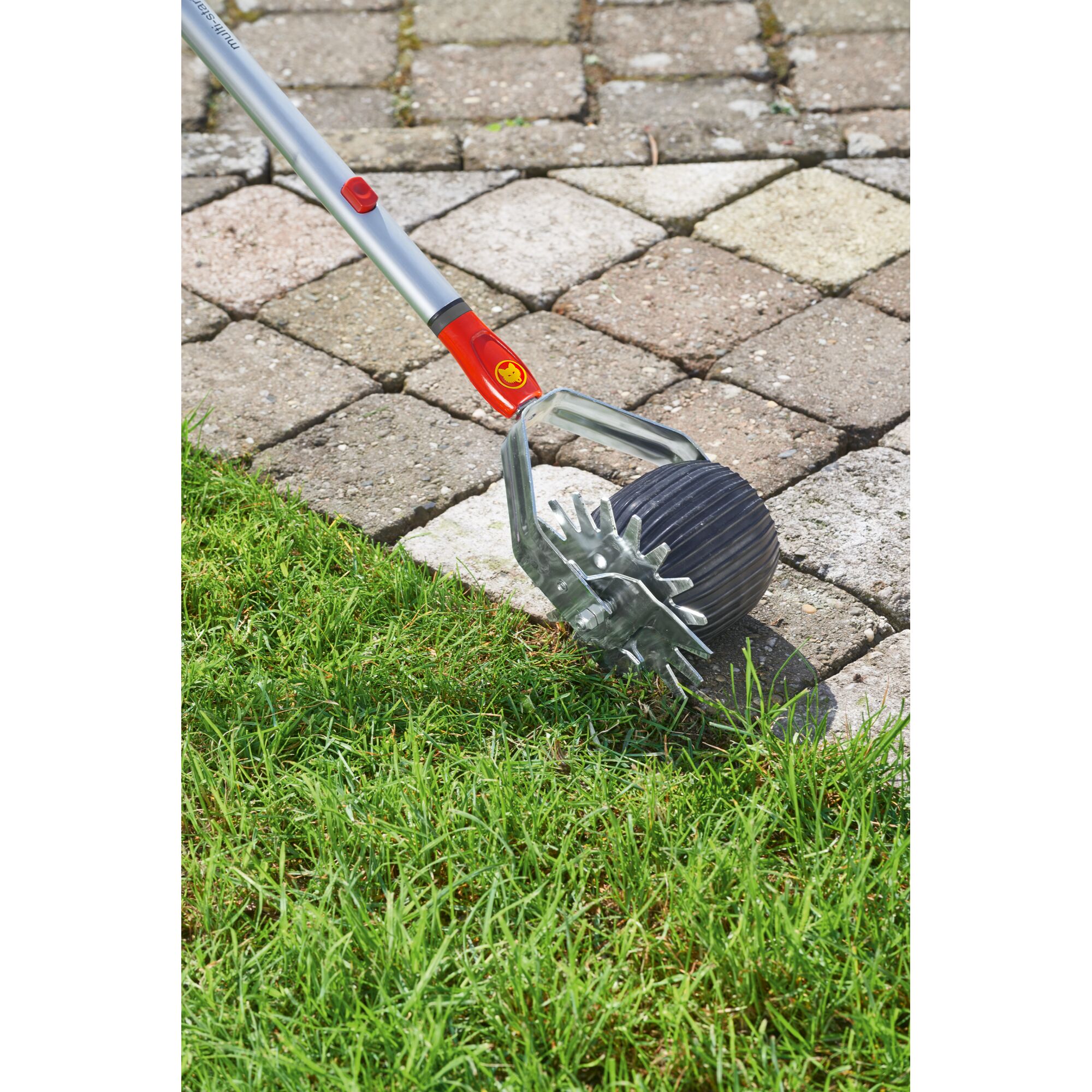 A metal and plastic garden tool with a red and yellow handle is shown being used to edge grass along a stone walkway.