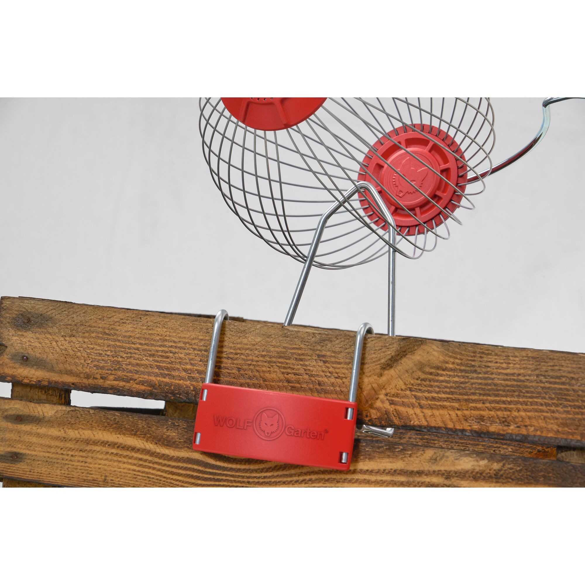 A close-up of a WOLFGarten garden tool with a red branded attachment and metal wire basket, placed on a wooden crate.