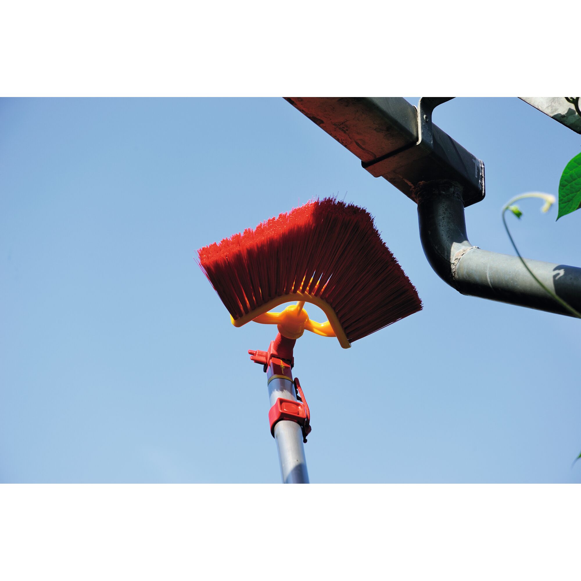 A red angled outdoor broom attached to a telescopic handle is being used to clean a house gutter under a blue sky.