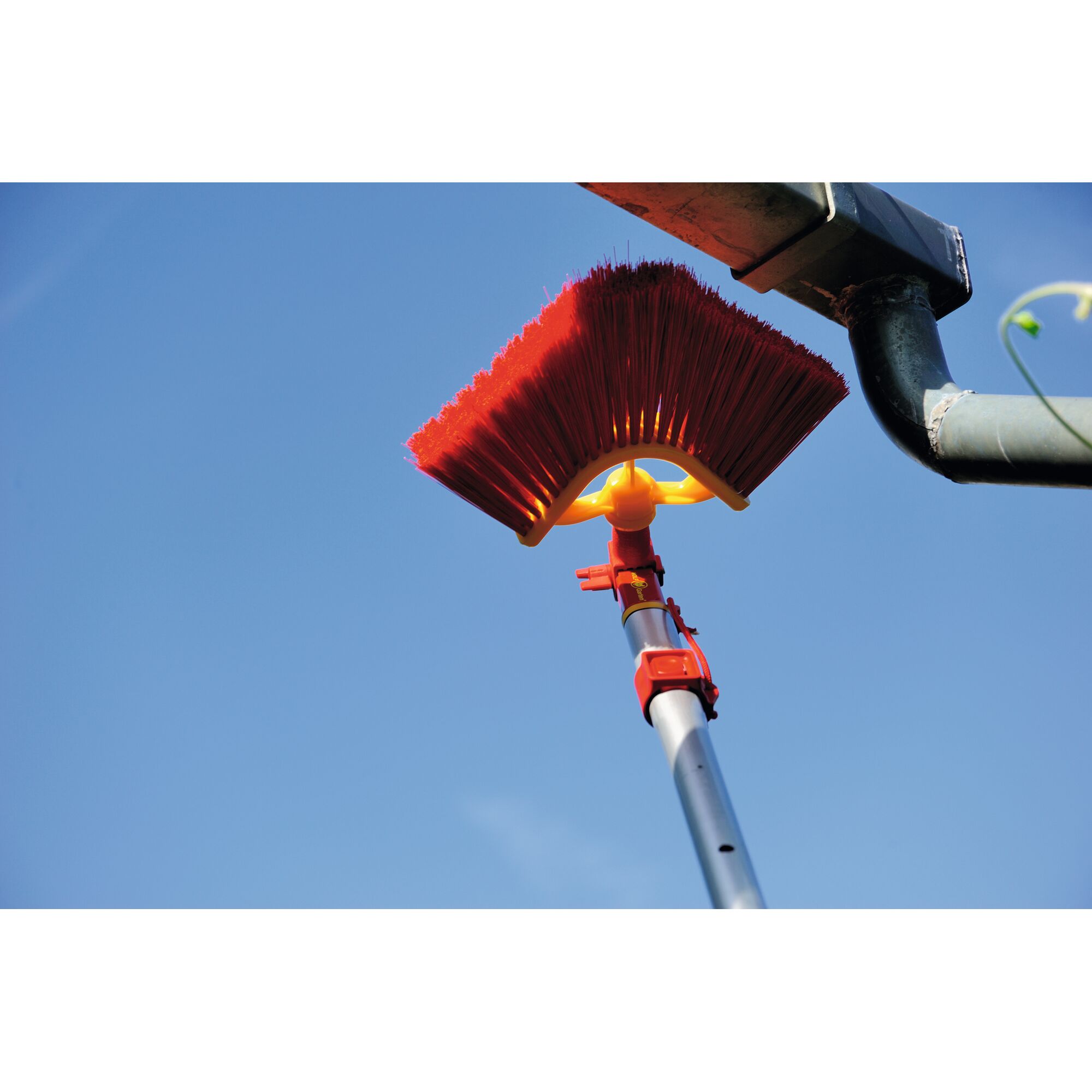 A close-up of a red angled broom attached to a telescopic handle, being used to clean a house gutter against a clear blue sky.