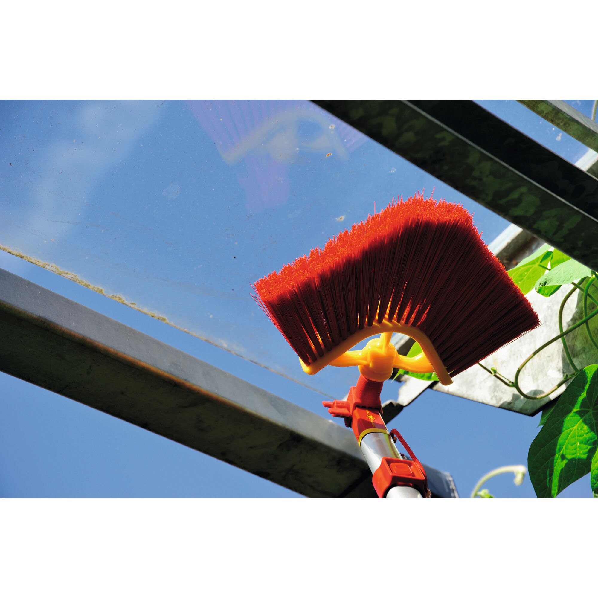 A close-up view of a red angled broom with a yellow handle attachment being used to clean the glass roof of a greenhouse, with a blue sky visible in the background.