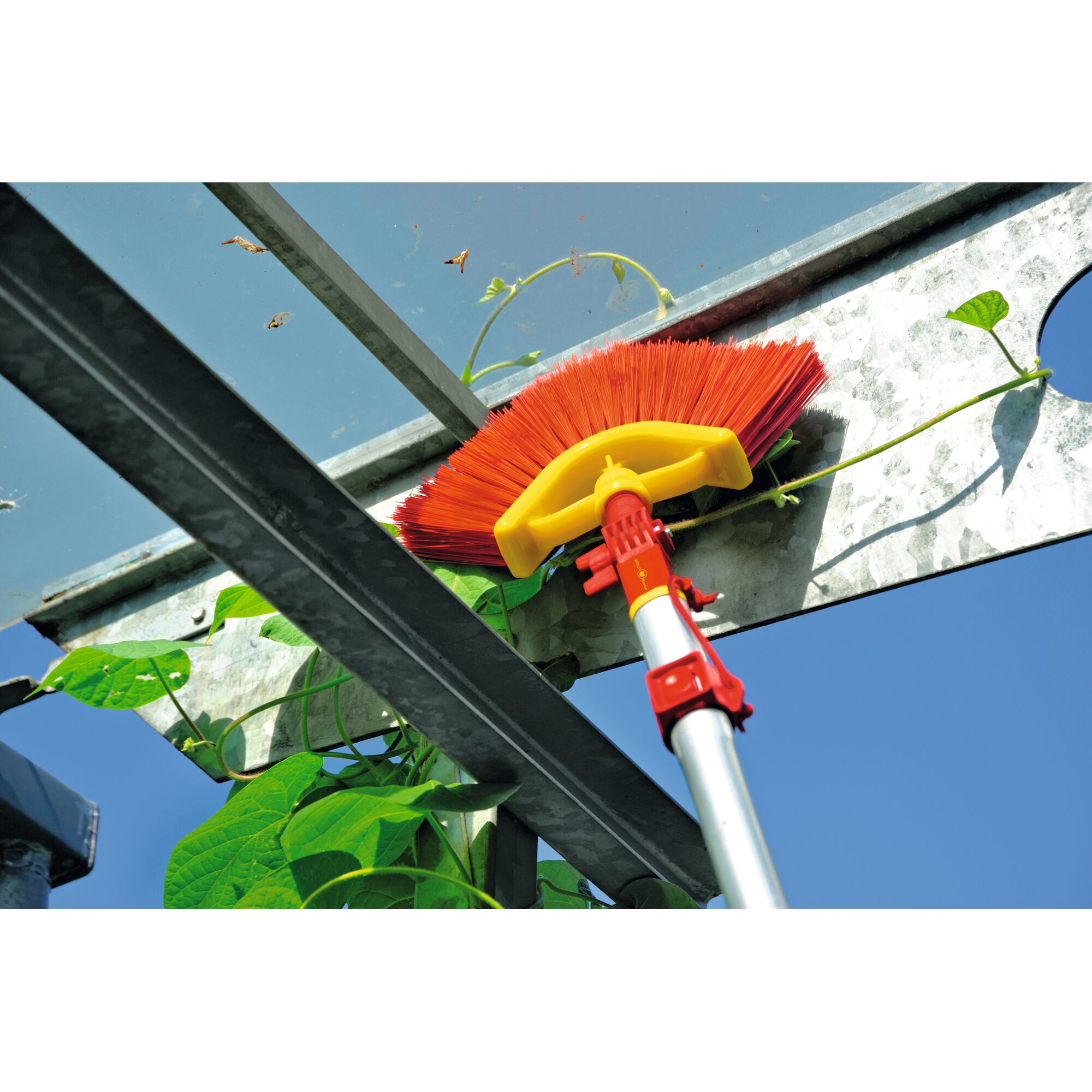 A close-up view of a red and yellow telescopic brush cleaning the upper metal structure of a greenhouse with green plants and blue sky visible in the background.