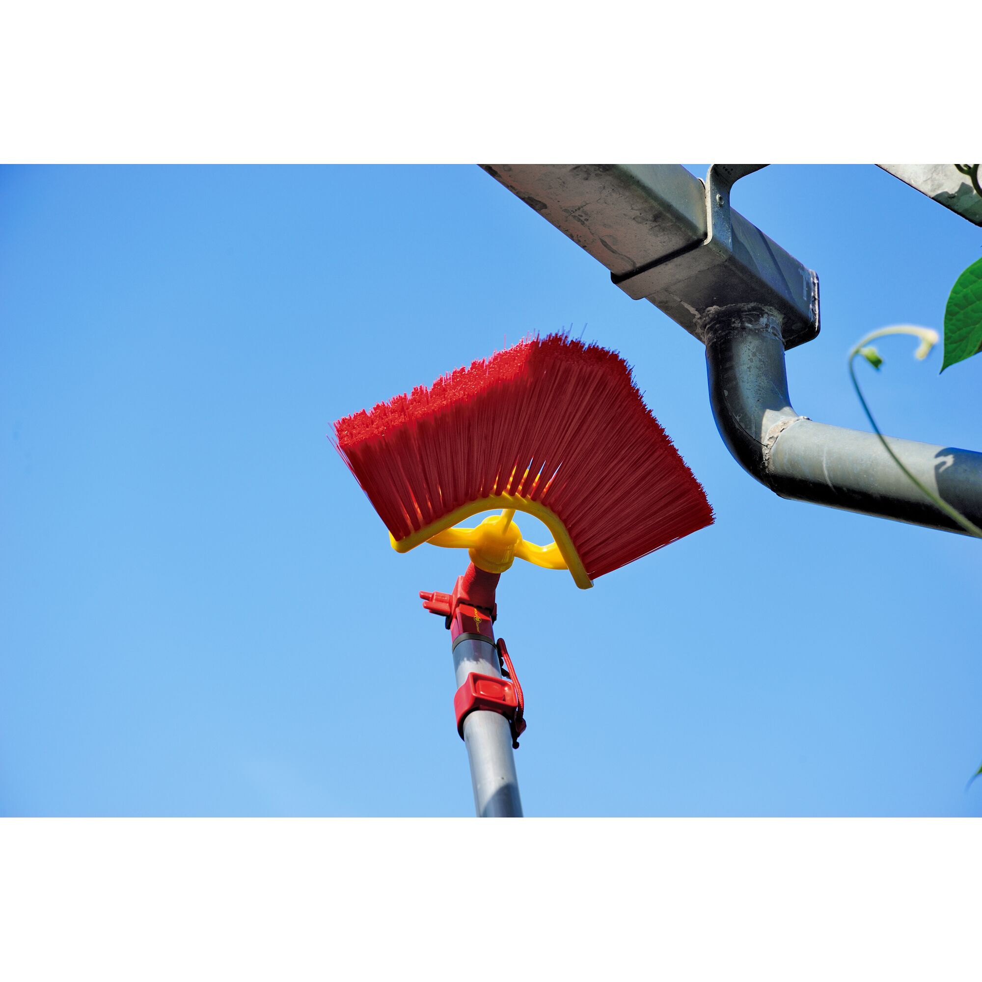 A red and yellow angled brush attached to a telescopic pole, being used to clean a metal rain gutter against a blue sky.