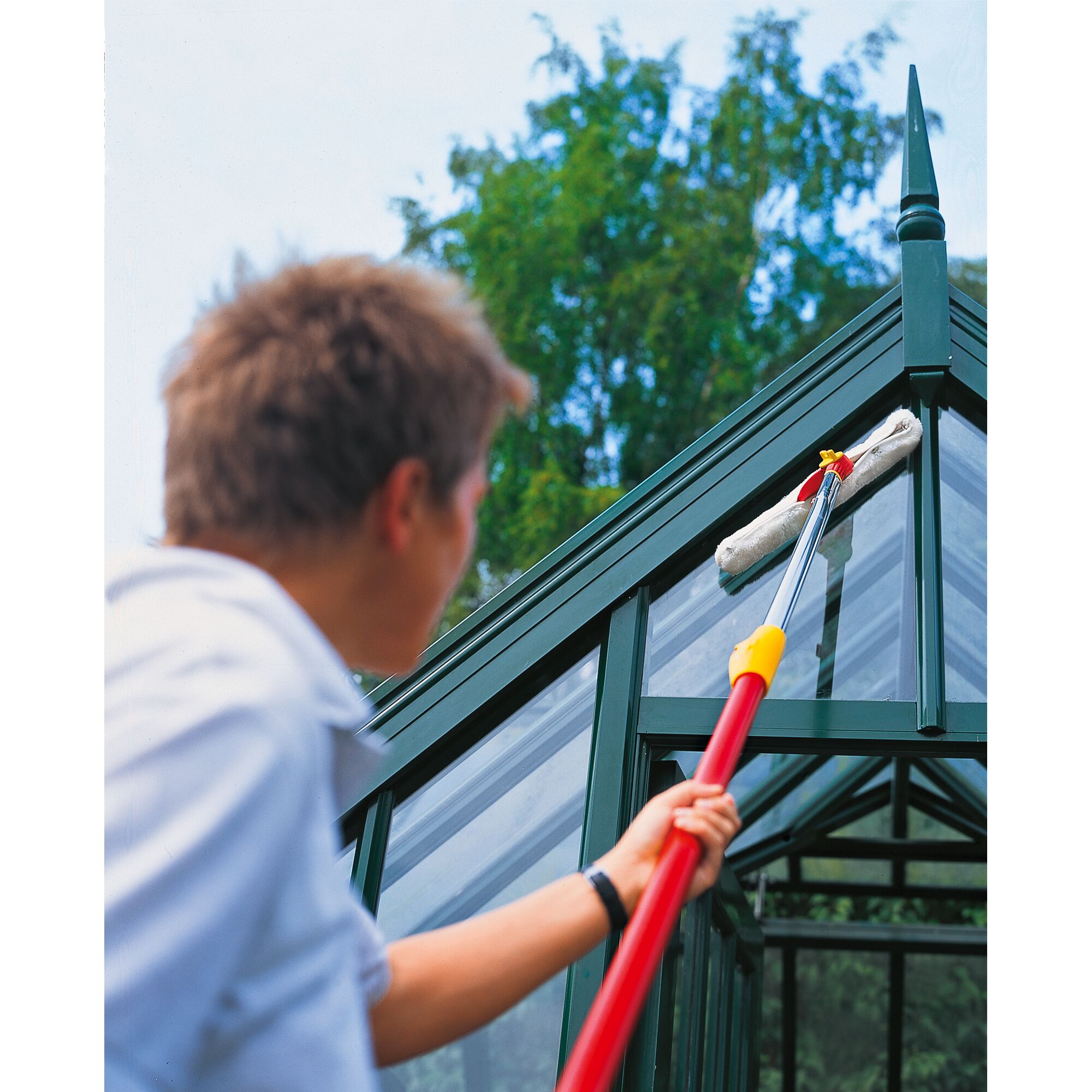 Person using a WOLFGarten cleaning tool with a long handle to clean the glass roof of a greenhouse.