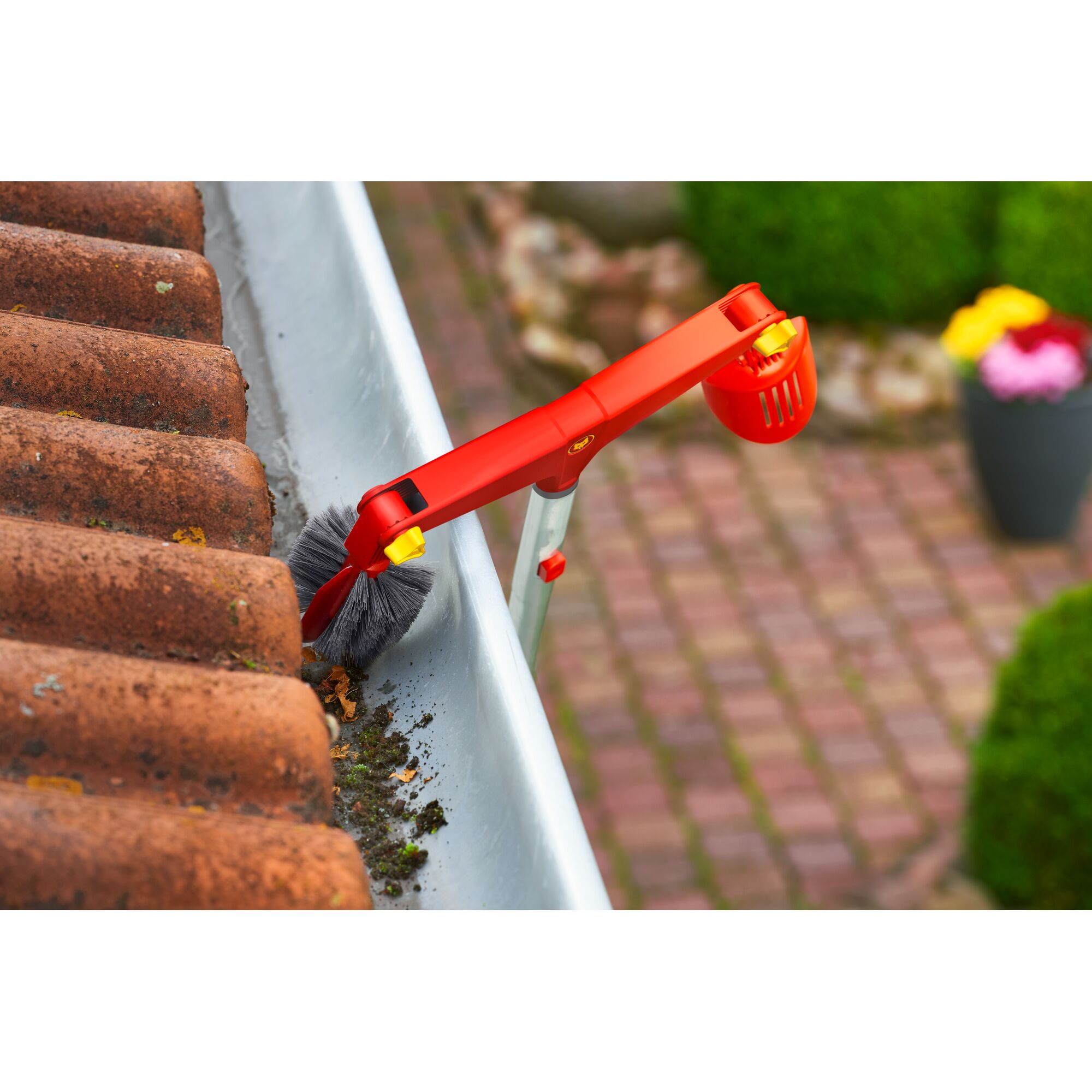 A red and yellow gutter cleaning tool with a brush attachment is being used to remove debris and dirt from a roof gutter next to terracotta roof tiles. The tool is attached to a telescopic handle, and there is a garden with a potted plant visible in the background.