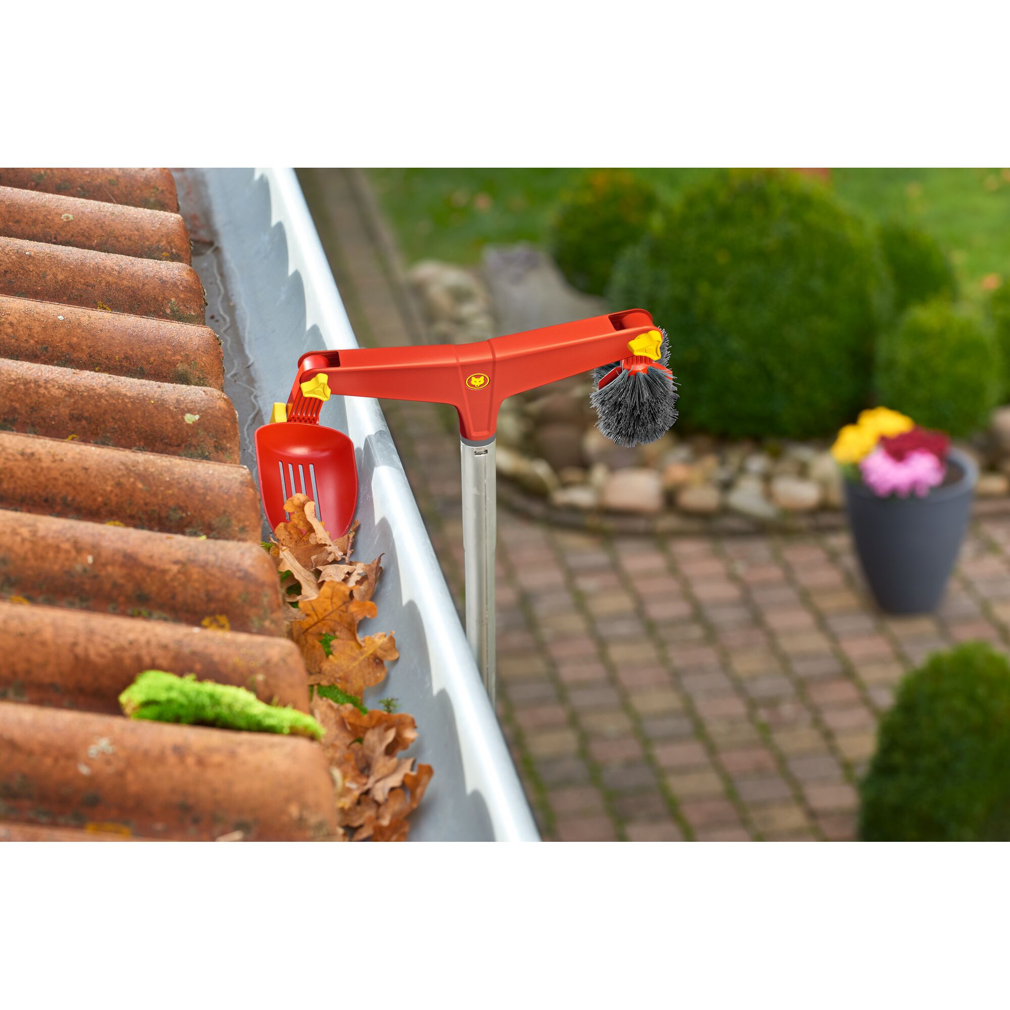 A red WOLFGarten gutter cleaning tool is being used to remove leaves from a roof gutter. The tool has a brush on one side and a scoop on the other, attached to a long handle.