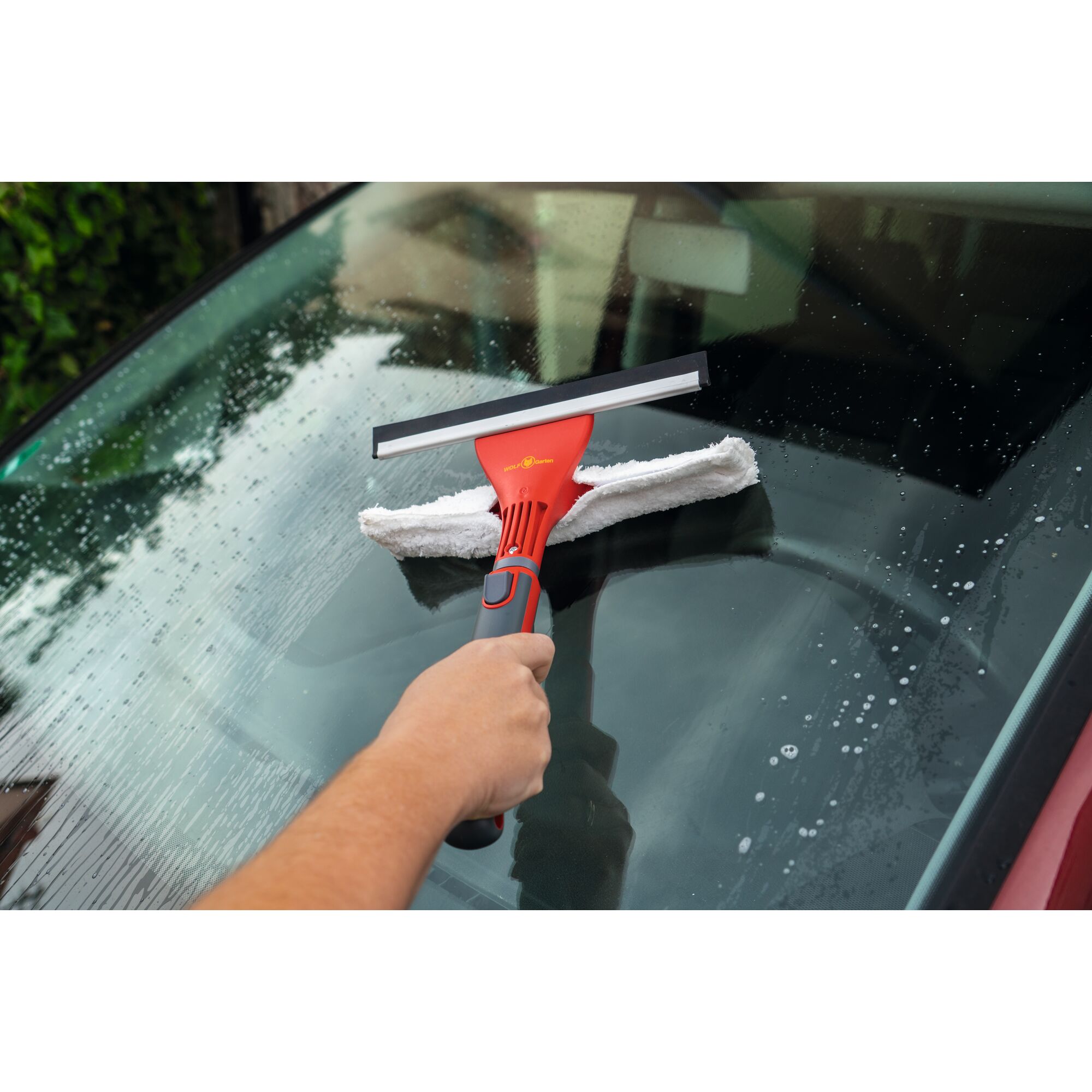 A hand is using a WOLFGarten tool with a red handle and a squeegee to clean the windshield of a car. The tool also has a white cleaning pad attached.