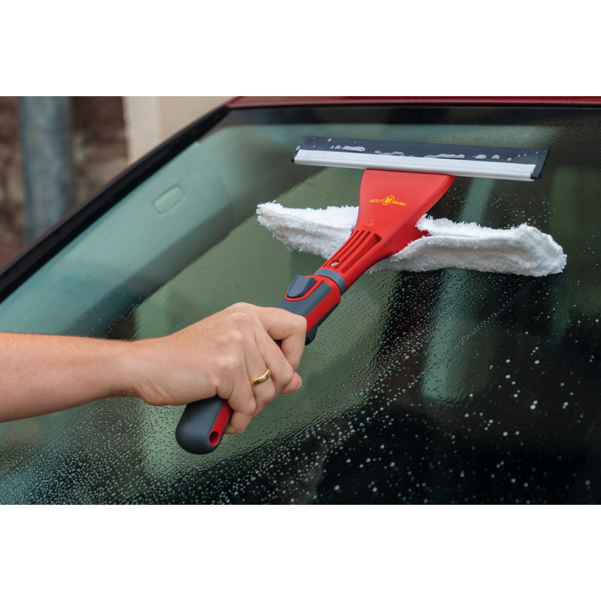 A hand holds a WOLFGarten window cleaning tool with a red handle, cleaning a wet car windshield. The tool has a squeegee and a white cleaning cloth attached.