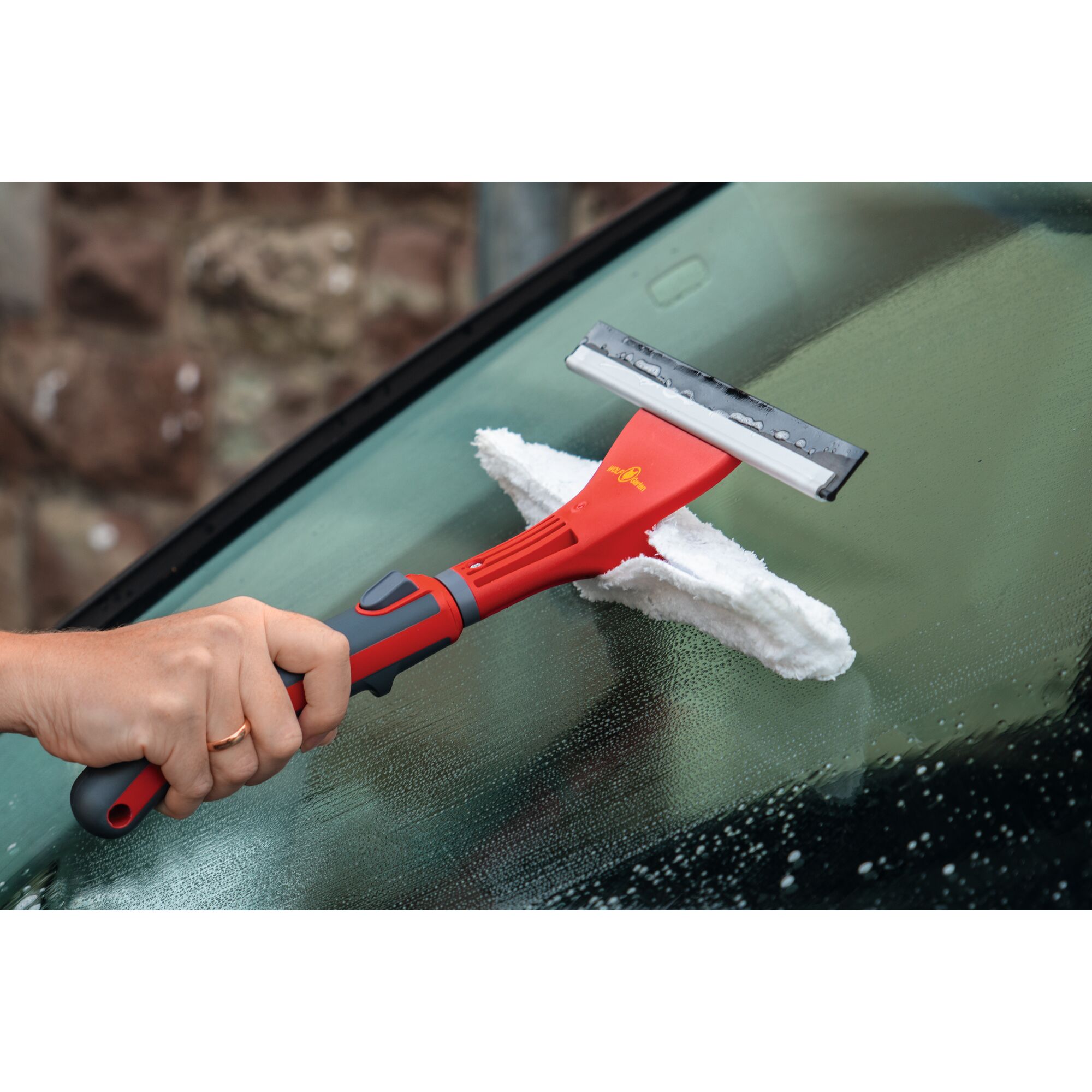A person using a WOLFGarten red and black window cleaning tool with a squeegee and microfiber pad to clean a wet car windshield.