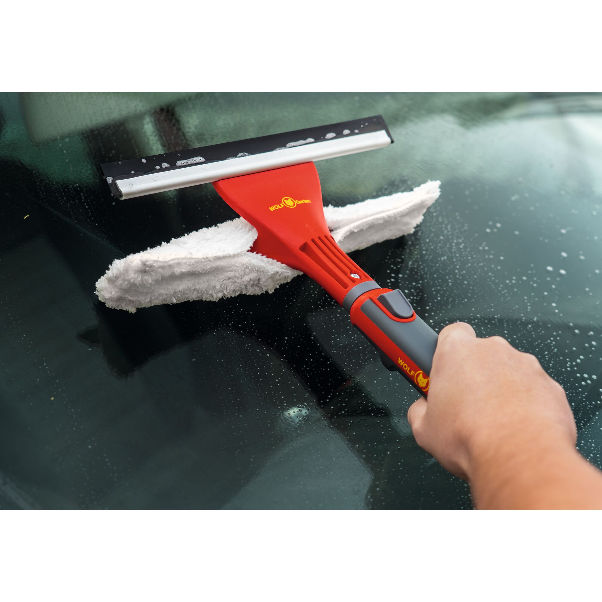 A person is using a WOLFGarten red-handled cleaning tool with a squeegee and microfiber pad to clean a glass surface with water droplets.