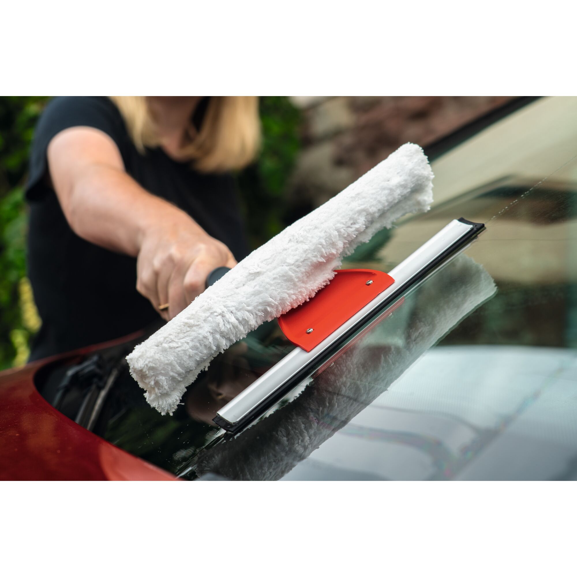 A person is cleaning a car windshield with a WOLFGarten tool that has a white cleaning pad and a red handle.