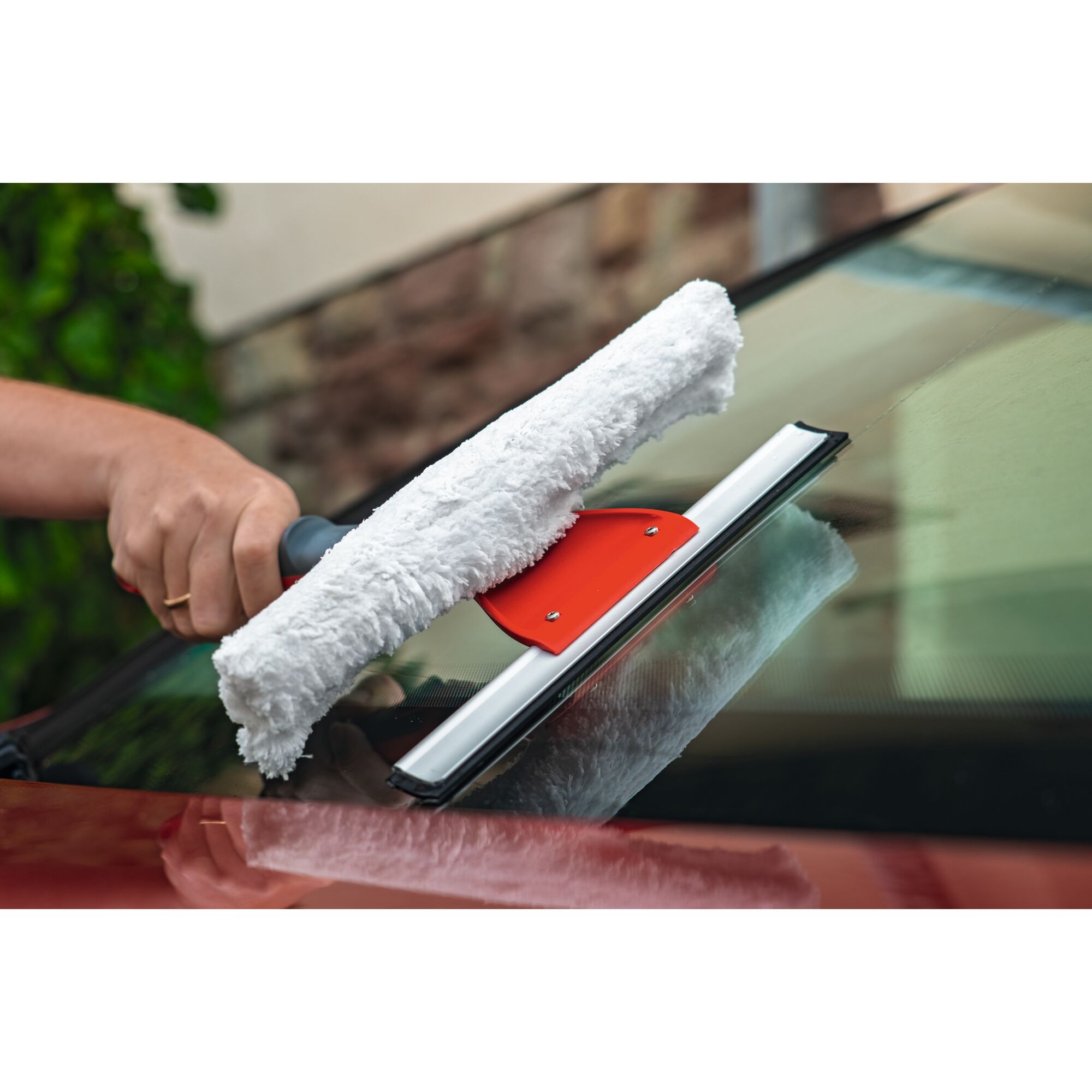 A person uses a cleaning tool with a white microfiber pad and a red plastic base to wash and wipe a car windshield.