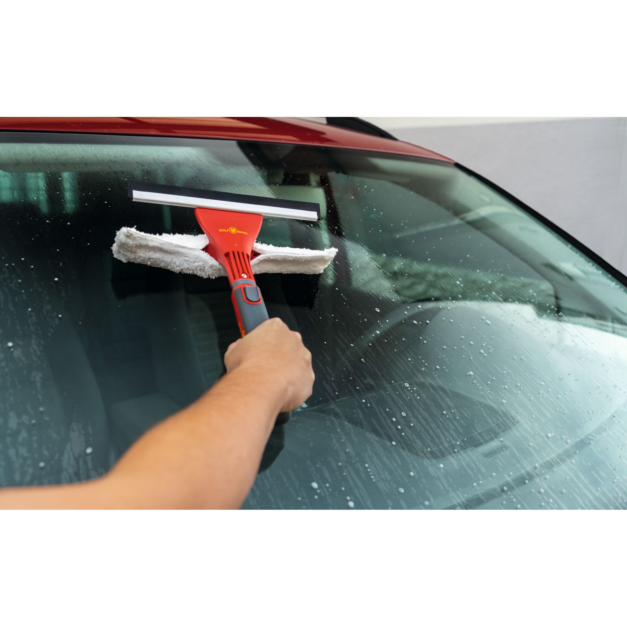 A person is using a WOLFGarten red-handled window cleaning tool with a squeegee and microfiber cloth to clean the windshield of a car.