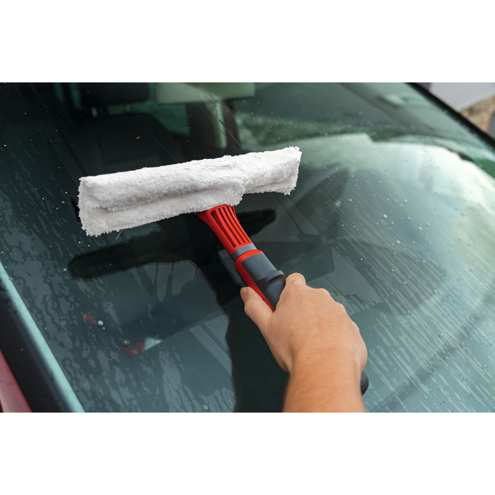 A person is cleaning a car windshield with a red and gray squeegee tool that has a white microfiber pad.