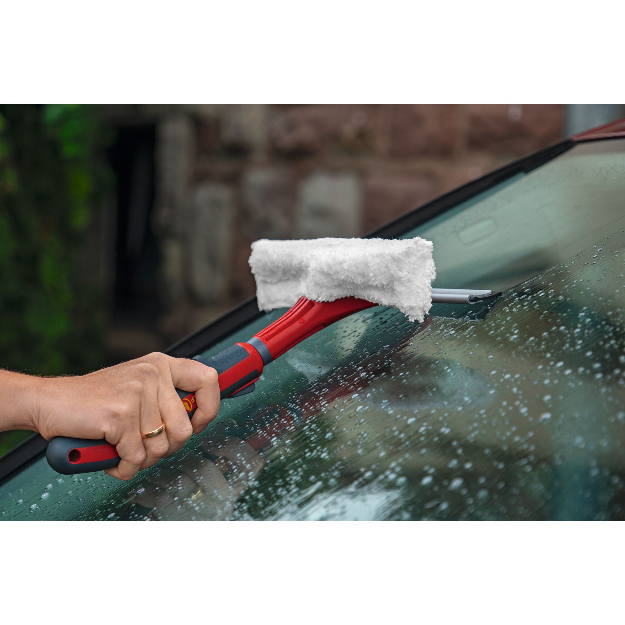 A person uses a red and gray window cleaning tool with a microfiber pad to clean a wet car windshield.