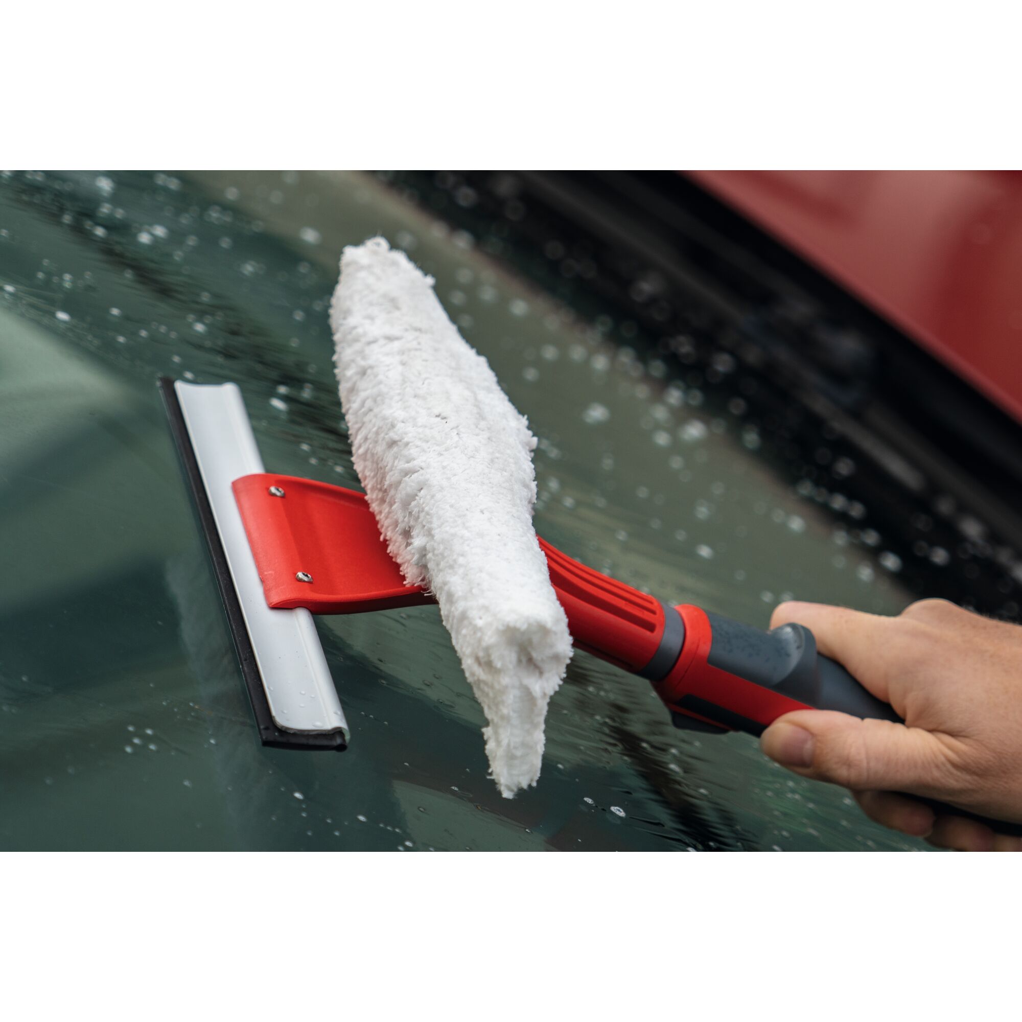 A person uses a red and black window cleaner tool with a white microfiber cloth to clean a wet car windshield.