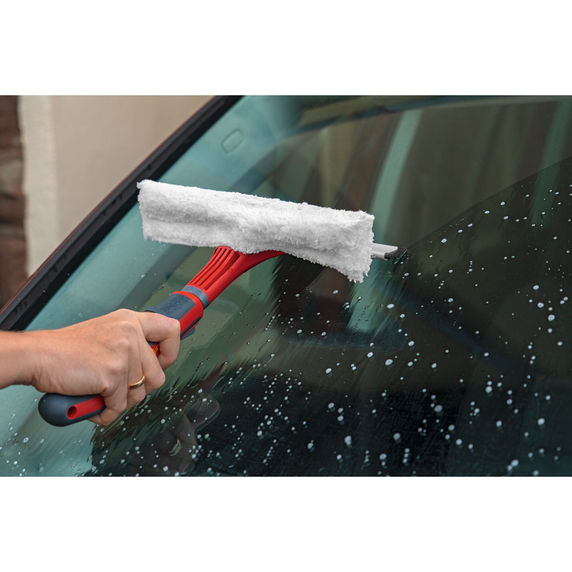 A person using a red and grey handled cleaning tool with a white microfiber pad to wash a car windshield with soap and water.