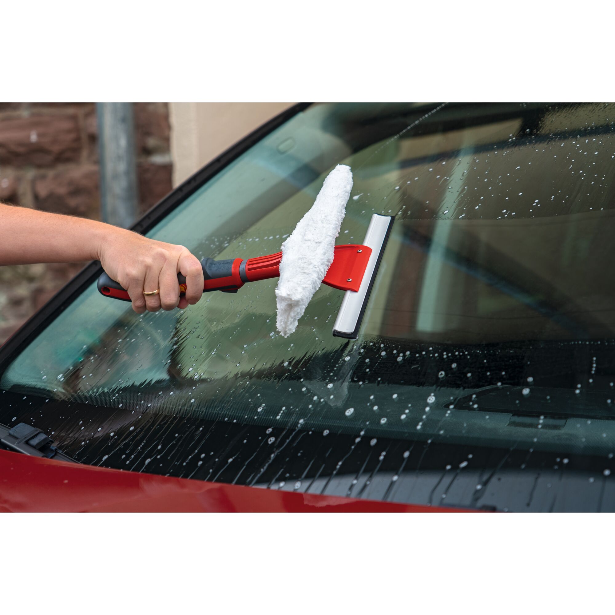 A person using a red-handled squeegee with a white sponge to clean a soapy car windshield.