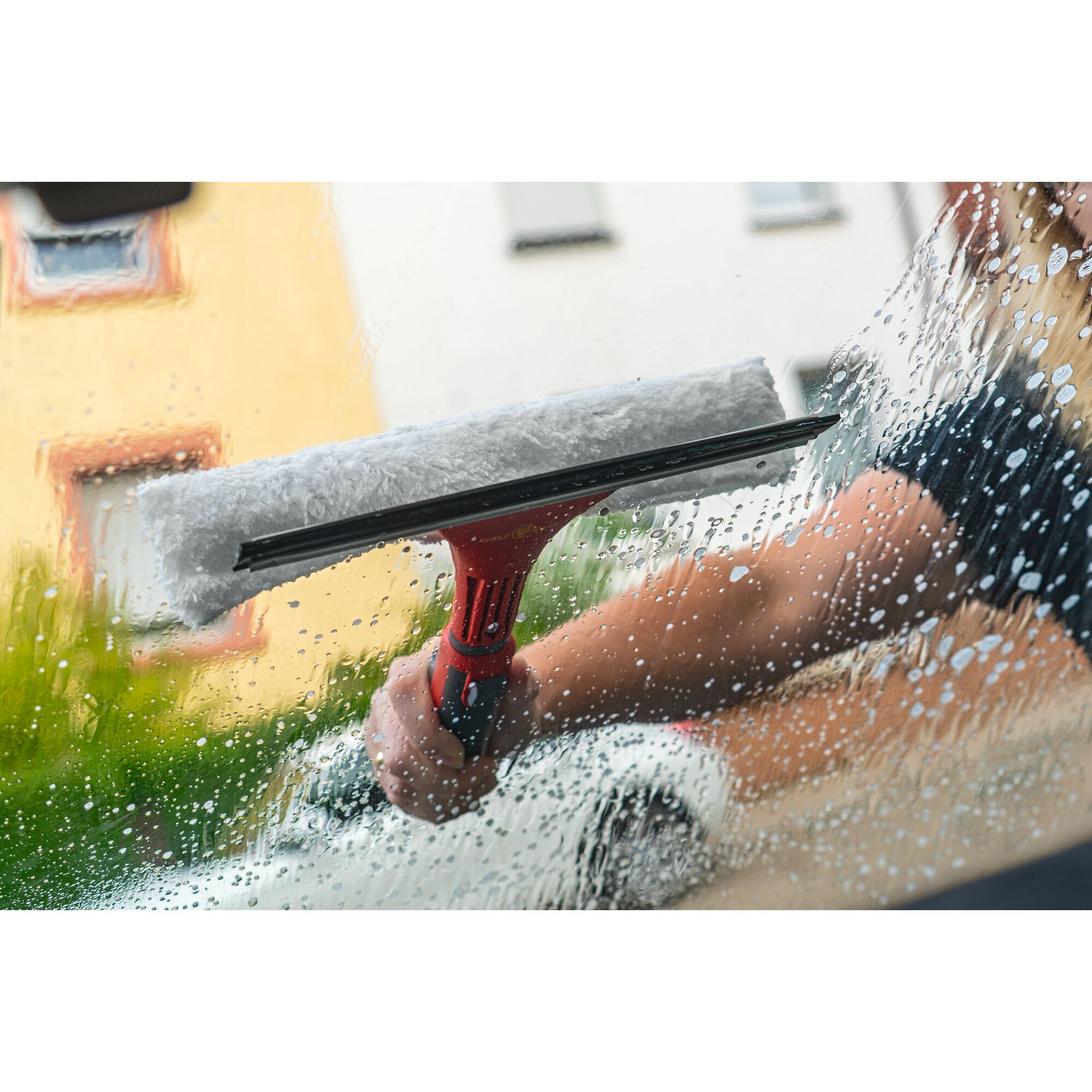 A person is cleaning a window with a WOLFGarten combination window washer and squeegee, featuring a red handle and soapy water.