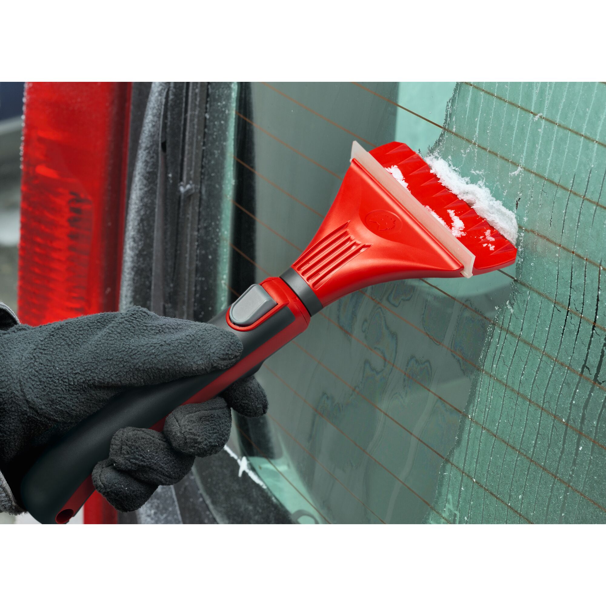A gloved hand uses a red and black ice scraper to remove ice from a car window.