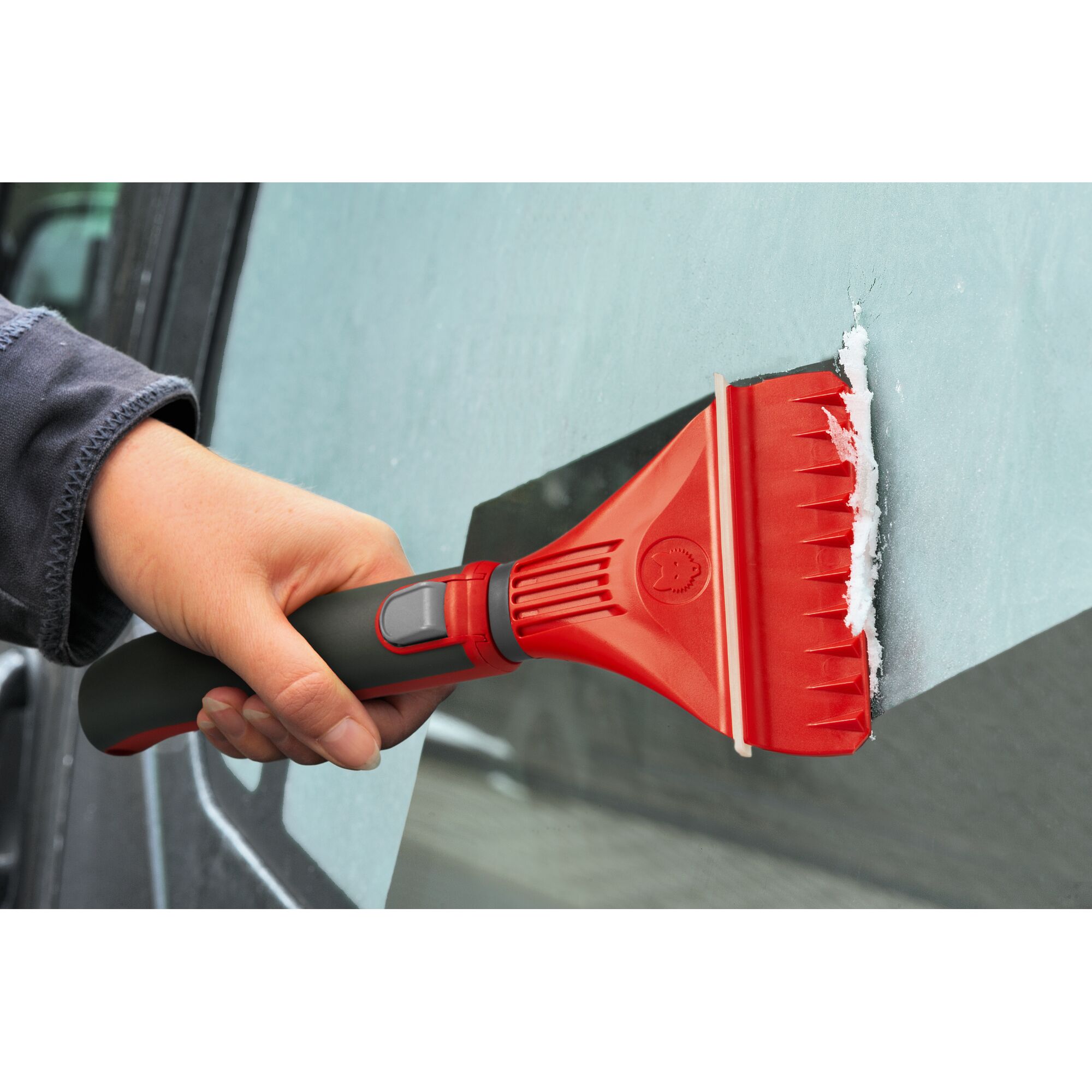 A person uses a red and black ice scraper to remove ice from a car window.