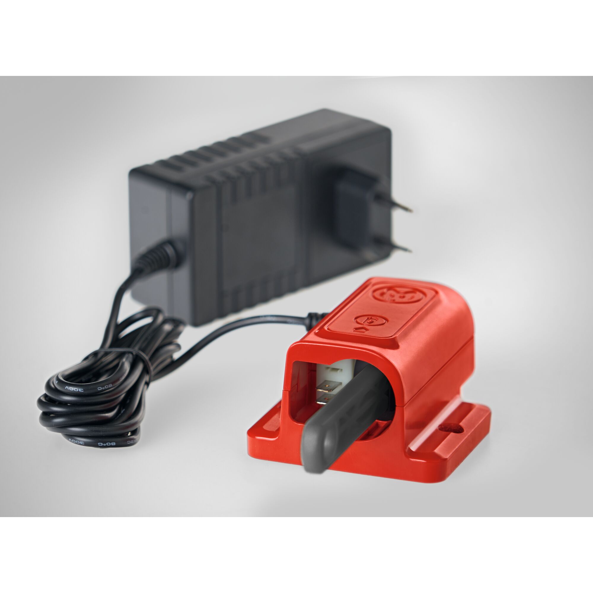A close-up image of an electronic battery charger with a red casing and a black power adapter. The charger has a slot for inserting a battery and is connected to the adapter with a cord. The background is neutral gray.