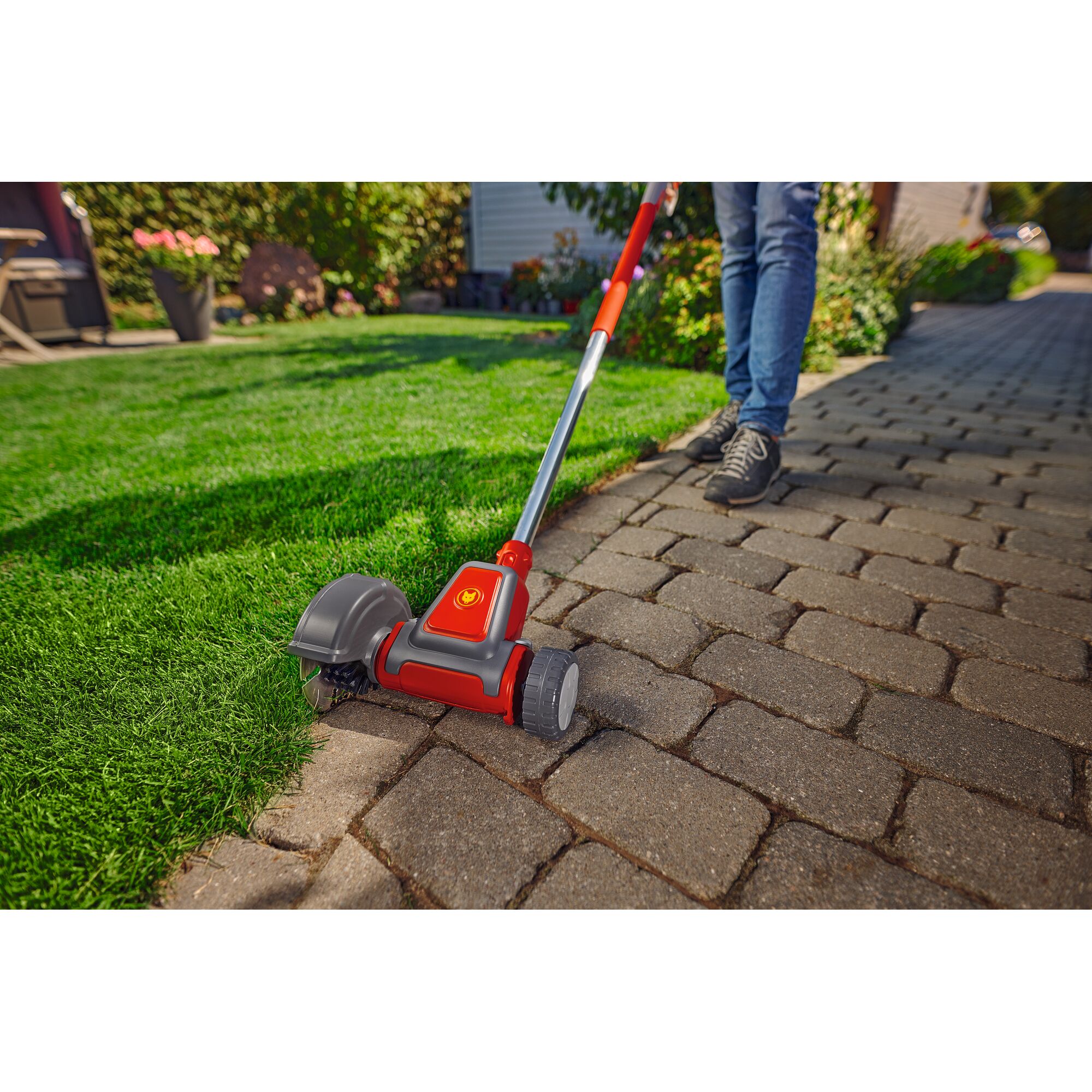 A person uses a red and grey WOLFGarten electric lawn edger to trim the edge between a grass lawn and a stone walkway. The tool is held by the handle, and the grass is neatly separated from the walkway.