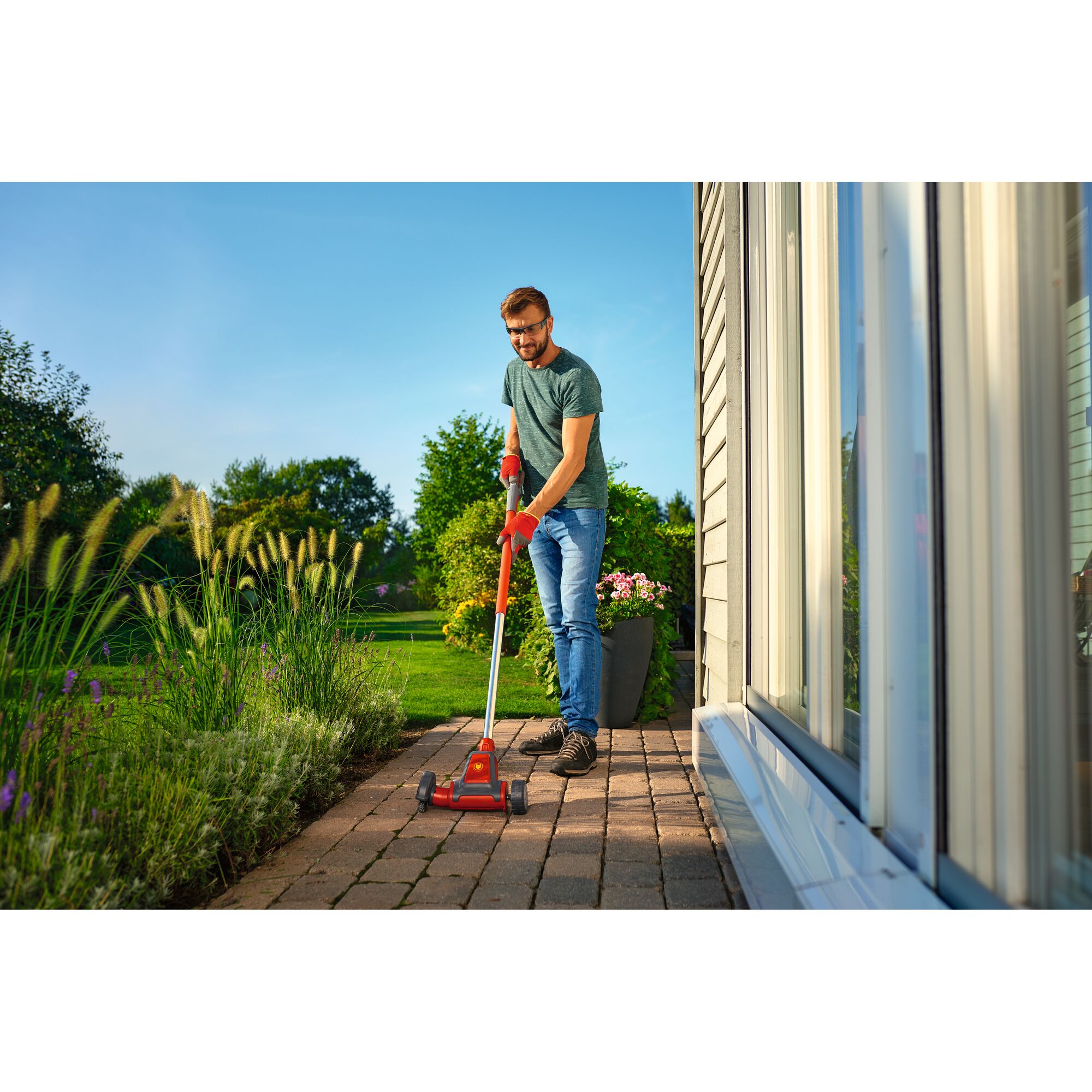 A person using a WOLFGarten garden tool with a long handle and wheels to clean between pavers on a patio next to a house. The person is wearing gloves and working outdoors beside a garden with plants and grass.