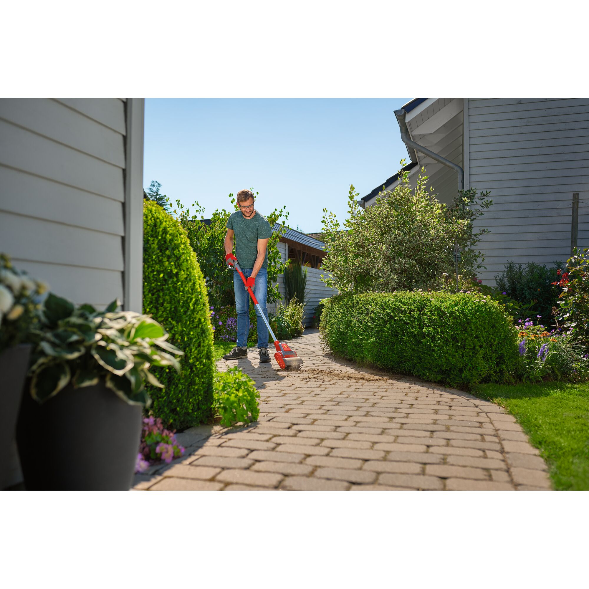 A person using a WOLFGarten garden tool to clean a paved pathway surrounded by green bushes and flowers in a residential garden.