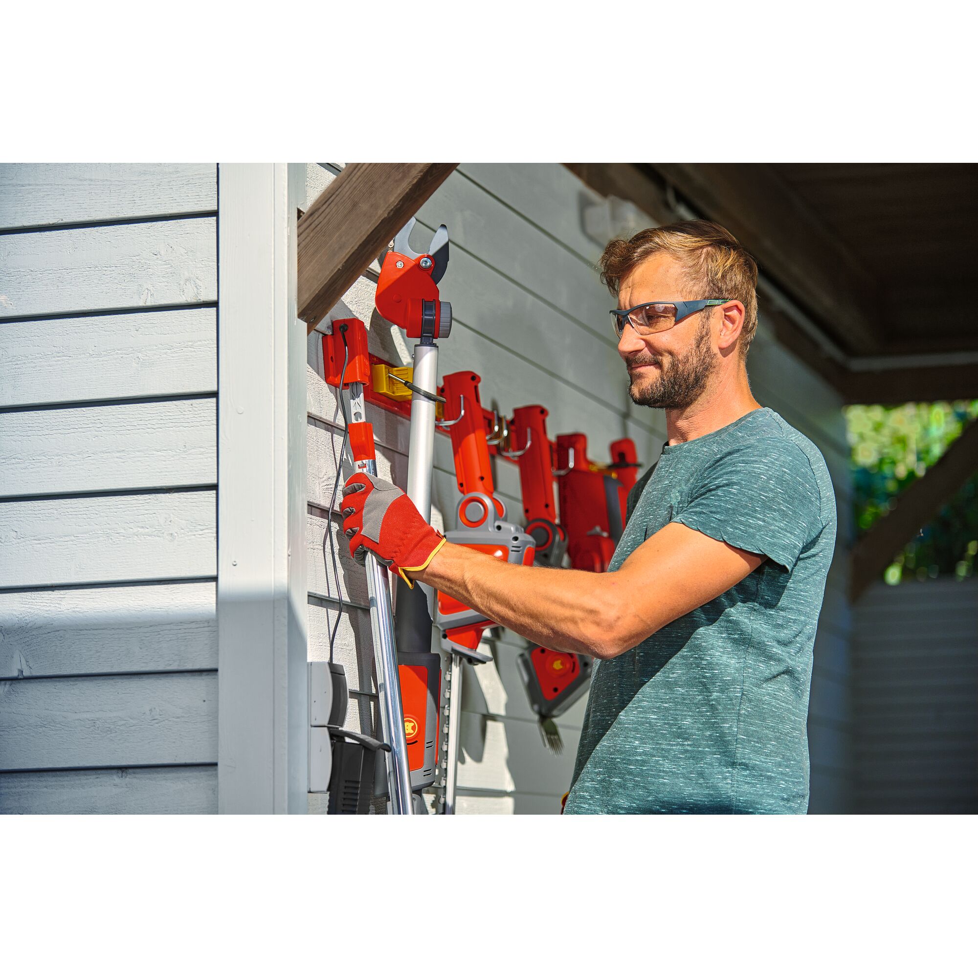 A person wearing red gloves is organizing red and silver garden tools, including pruning shears and other attachments, on a wall-mounted storage rack outside near a white wooden shed.