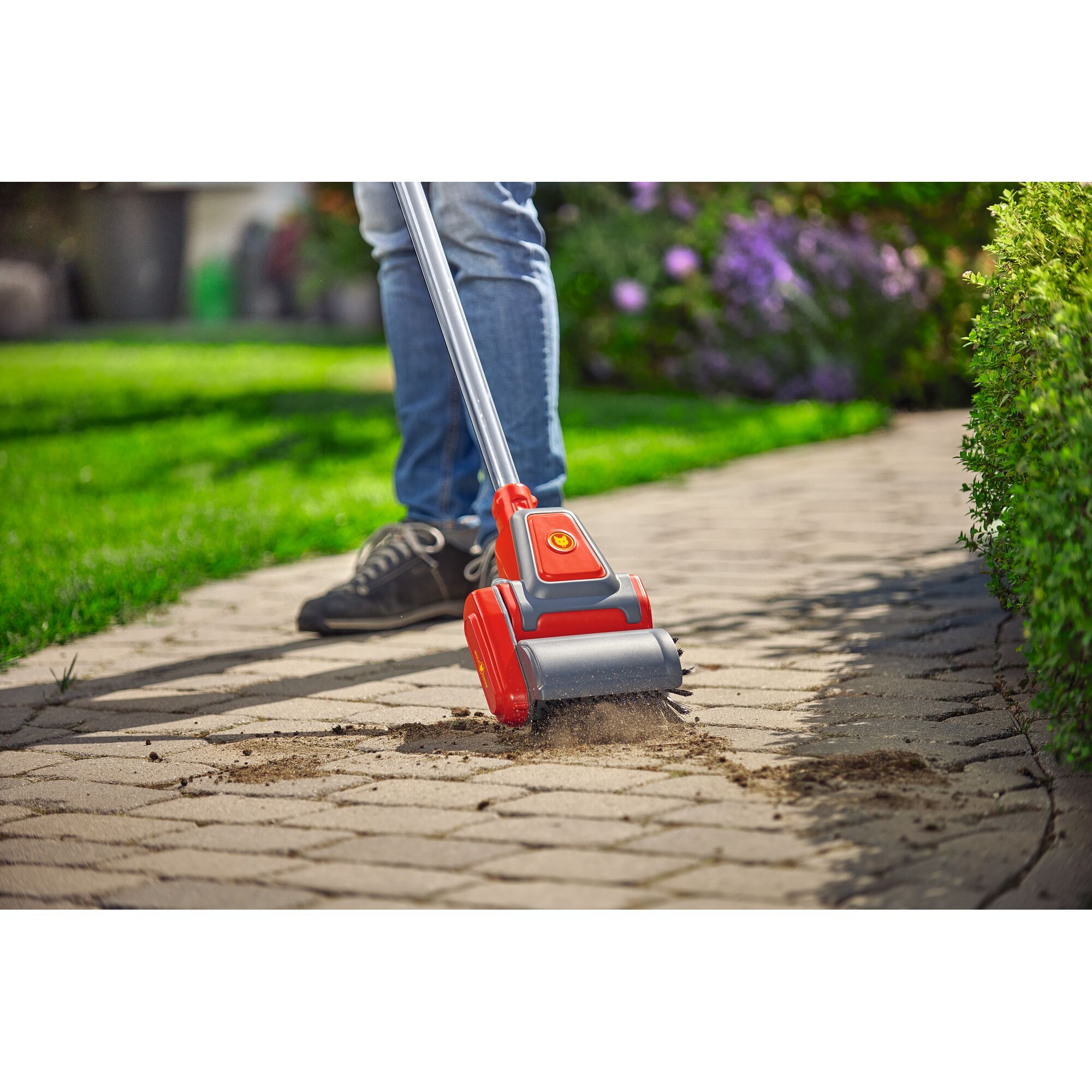 A person uses a WOLFGarten garden tool with a red and gray brush head to clean dirt from the gaps between paving stones on a garden pathway.