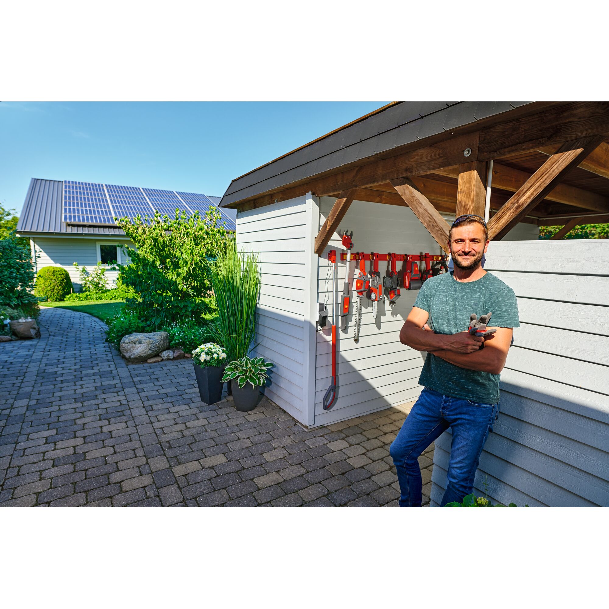 A person stands outside a wooden shed with gardening tools hanging on the wall, next to potted plants. The background shows a garden path leading to a house with solar panels on the roof.
