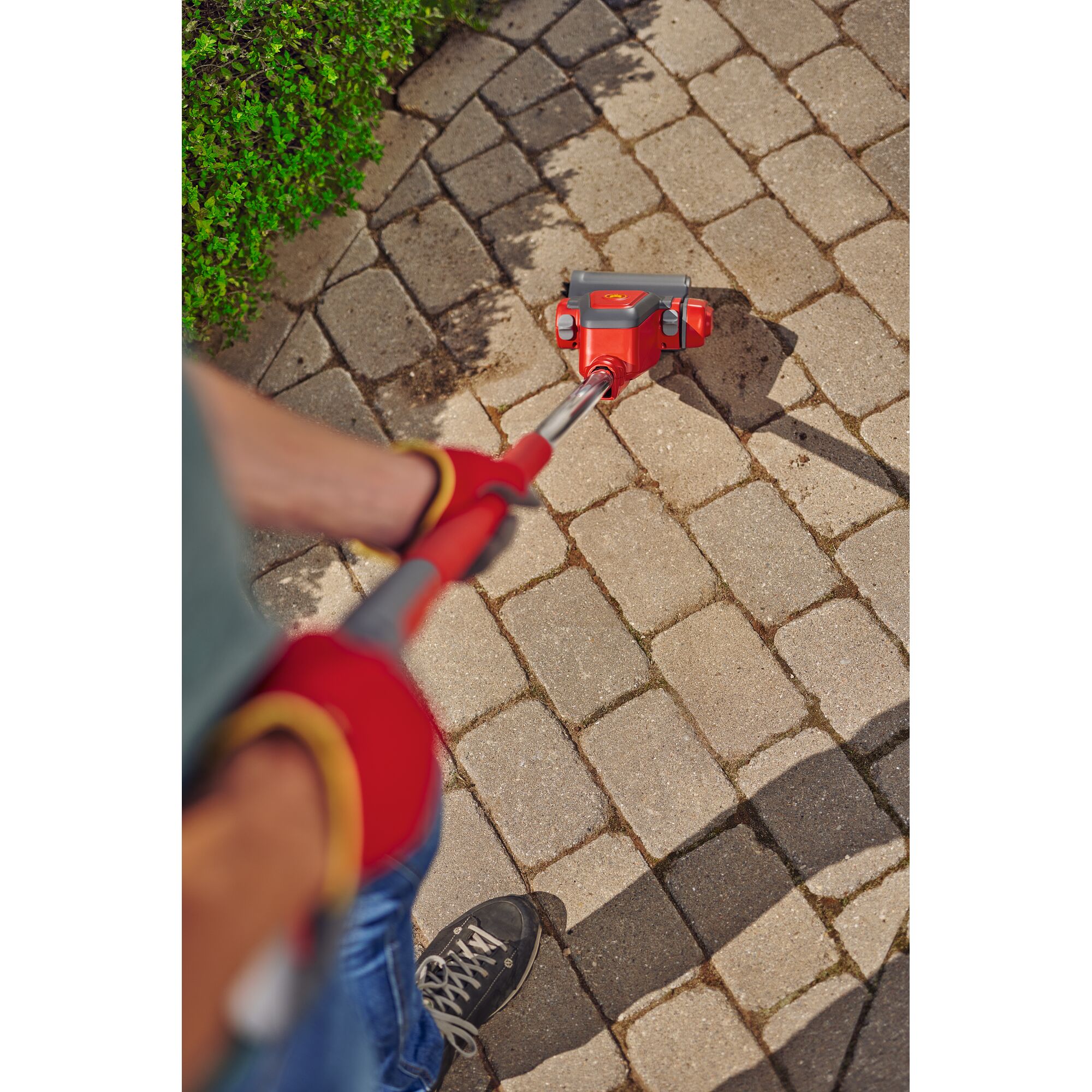 A person is using a red WOLFGarten tool to clean between the cracks of a stone pavement, wearing red gloves. Green bushes are visible in the corner.