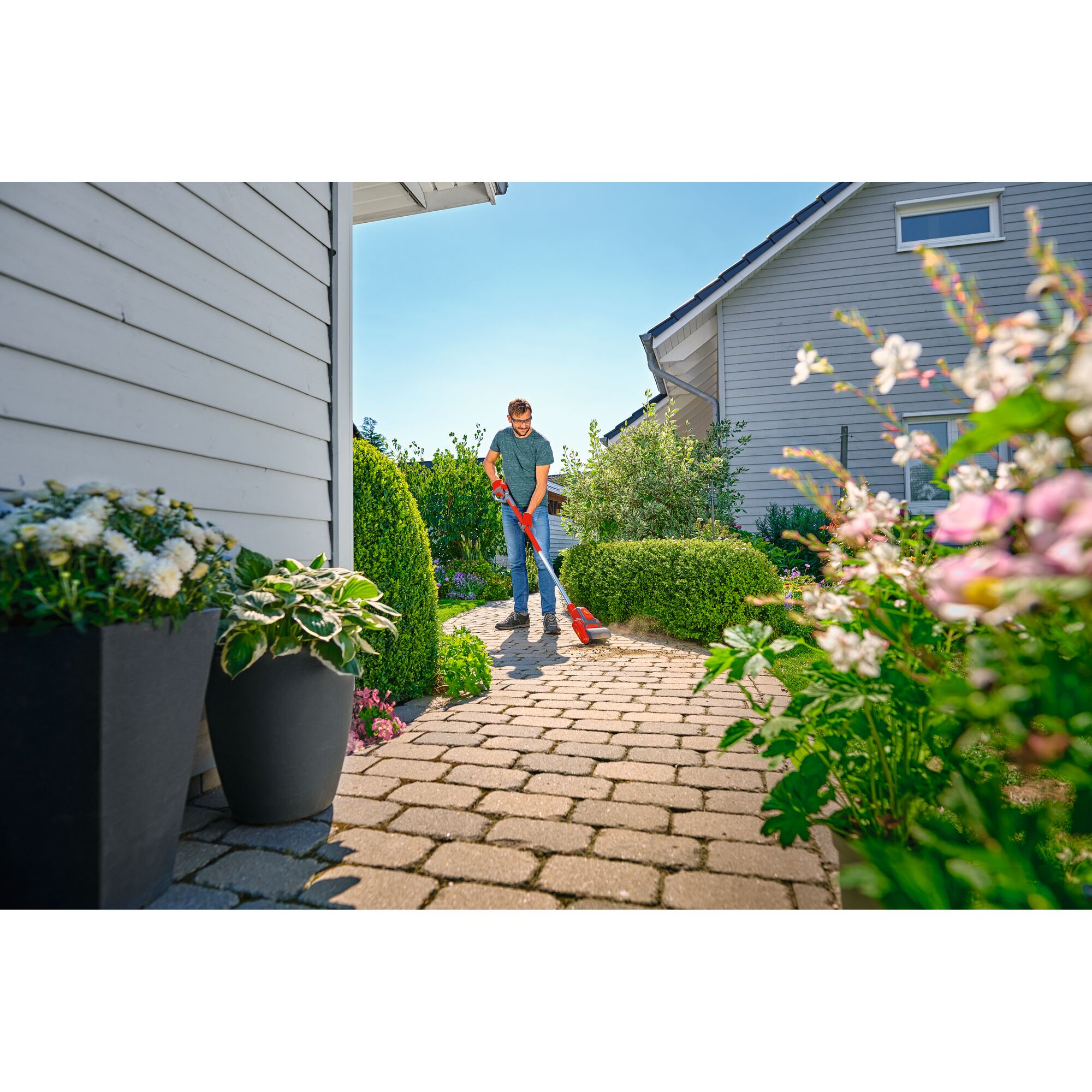 Person using a red WOLFGarten garden tool to clean a paved pathway surrounded by plants, bushes, and flowers in a residential garden area.
