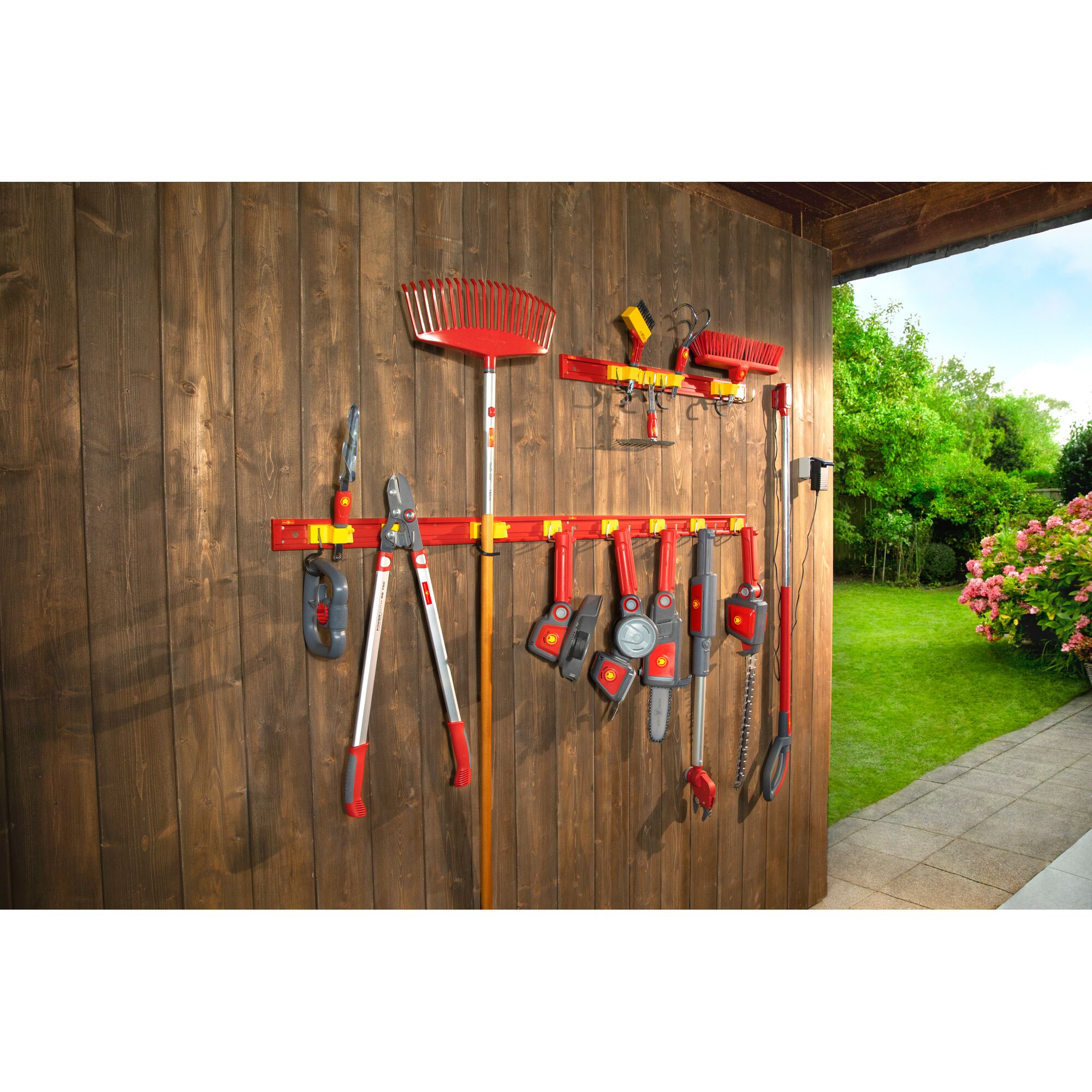 A variety of red and grey gardening tools and attachments are organized on a wooden wall-mounted rack. The rack is installed on an outdoor shed or wall, with a garden and greenery visible in the background.