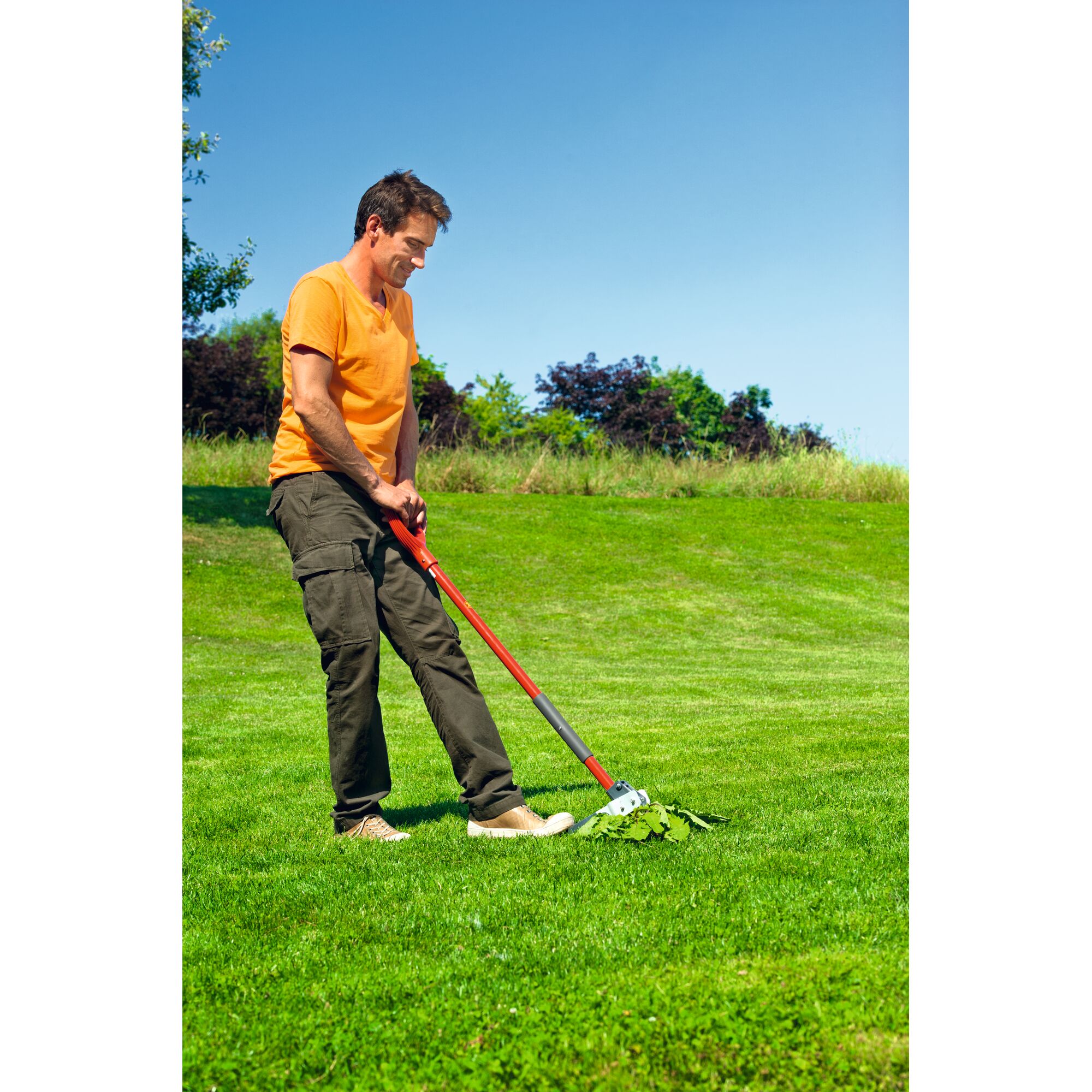 A person is using a red-handled garden rake to gather leaves on a green lawn, with trees and blue sky in the background.