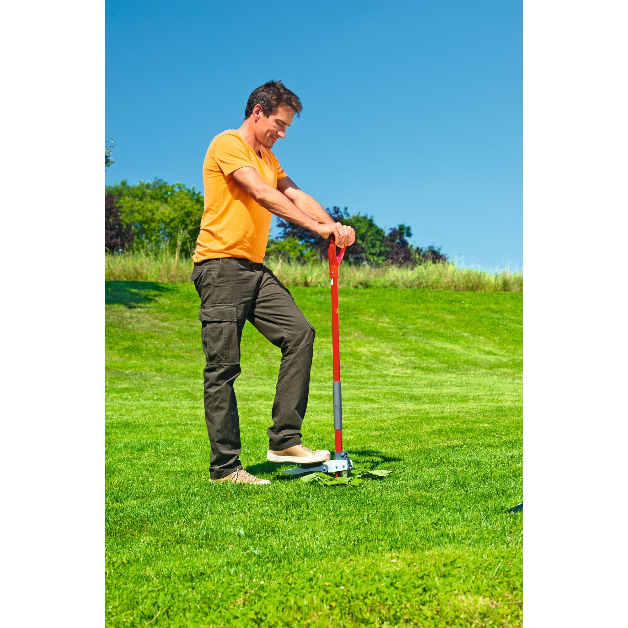 A person using a red-handled garden tool to remove weeds from a lawn, standing on green grass with a clear blue sky in the background.