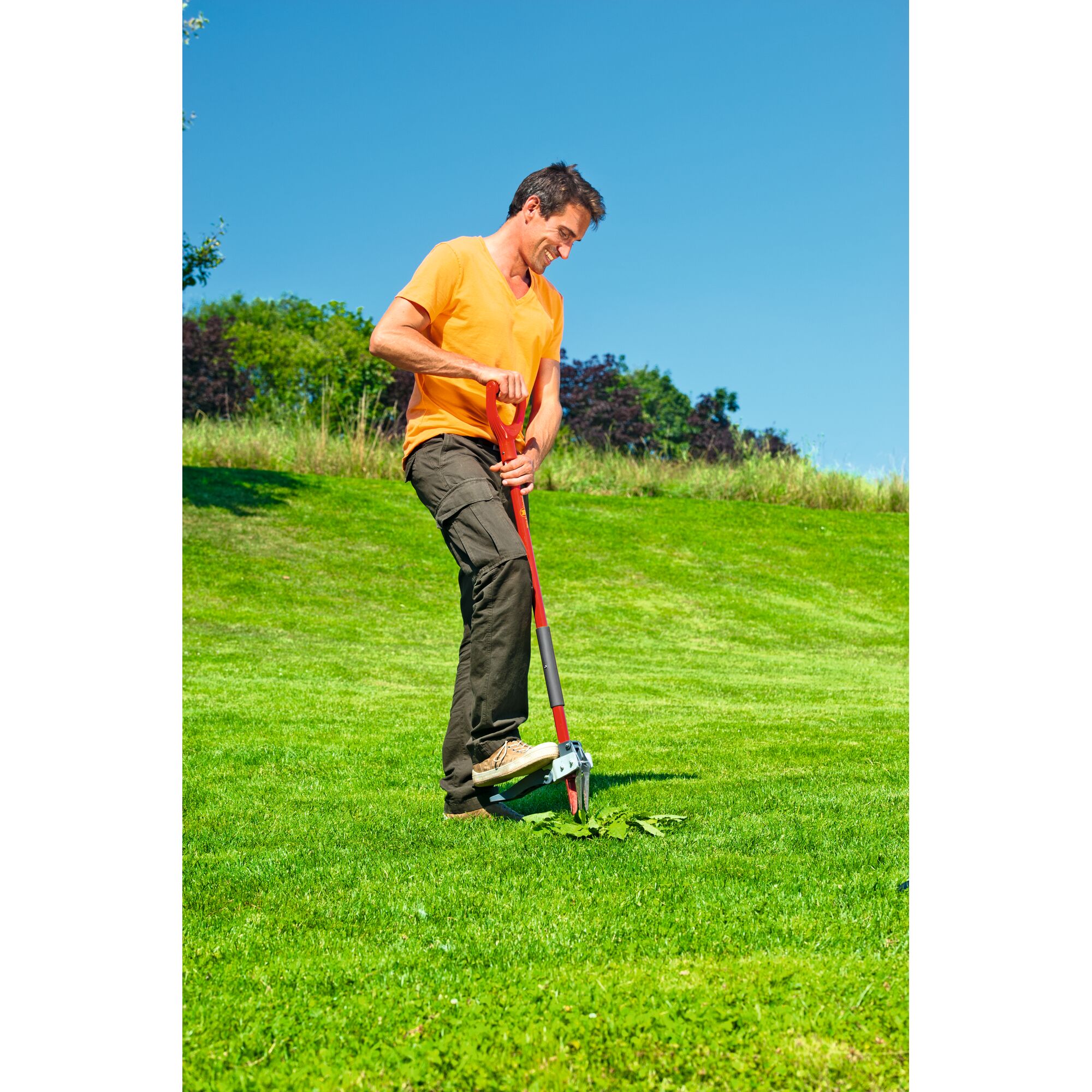 A person using a WOLFGarten garden tool to remove weeds from a lawn. The tool has a long red handle and is pressed into the ground with a foot pedal.