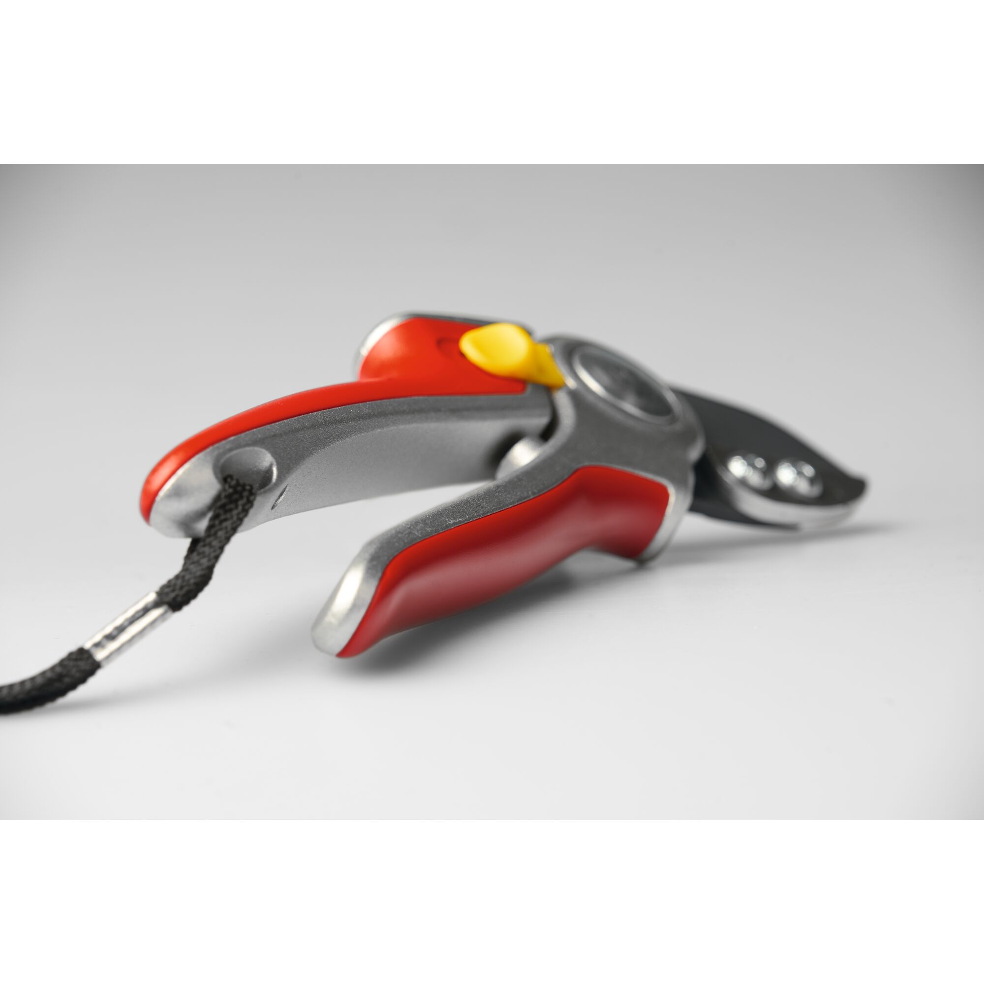 Close-up view of a red and silver garden pruning shear with a black cutting blade, yellow safety lock, and attached black wrist strap, placed on a neutral surface.