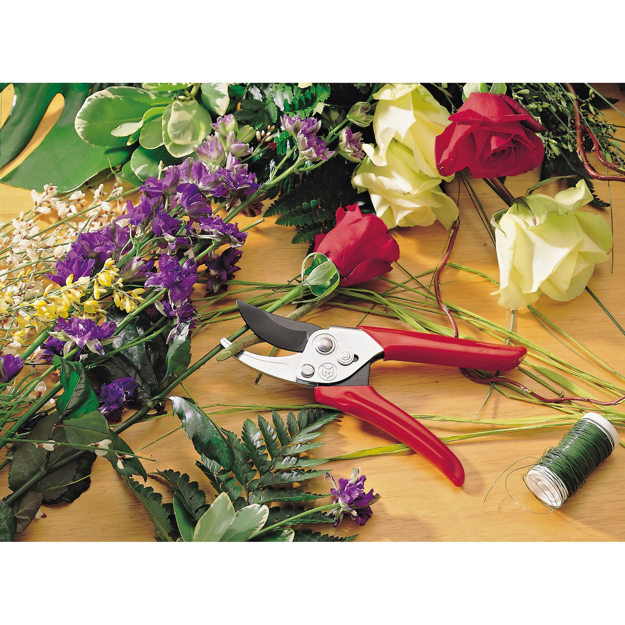 A pair of red-handled pruning shears is placed on a wooden table surrounded by various fresh cut flowers, including roses and purple blossoms, green leaves, and a spool of green wire. The scene suggests floral arrangement or gardening.