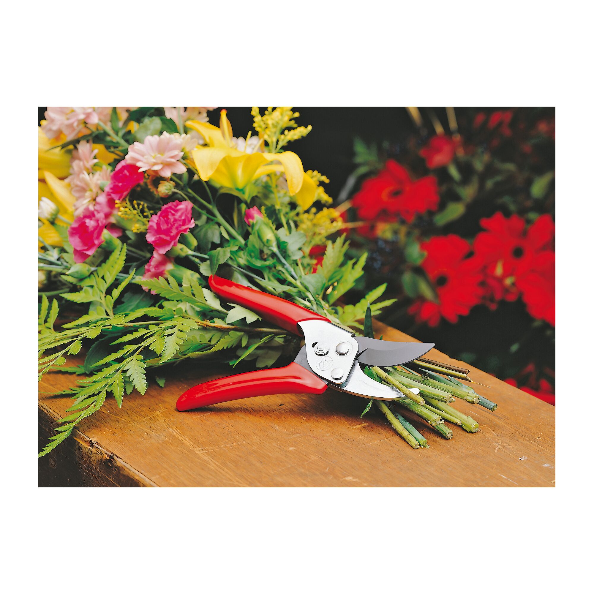 A pair of red-handled pruning shears rests on a wooden surface with a bunch of freshly cut colorful flowers and green foliage.