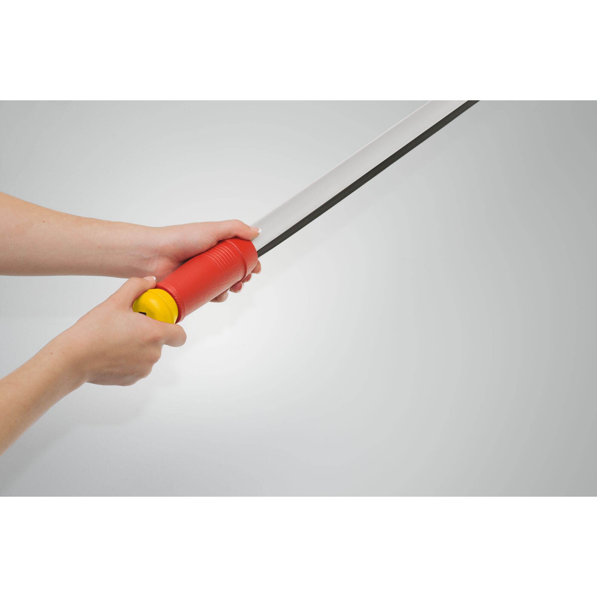 Close-up of hands holding a tool with a red handle and yellow end, featuring a long metal blade. The background is light gray.