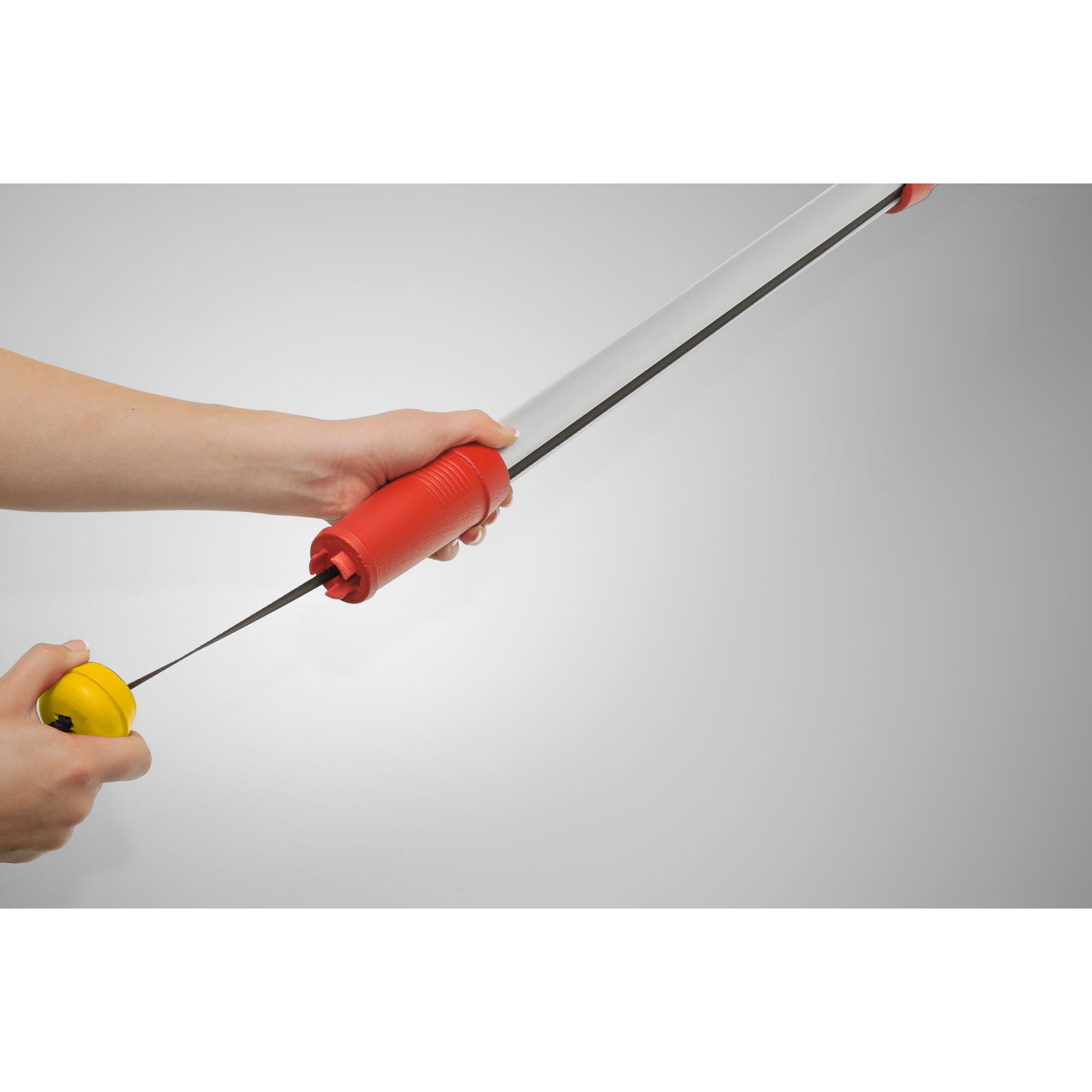 A close-up image of two hands holding a gardening tool with a long silver handle, a red grip, and a yellow circular component at the end. The tool appears to be used for pulling or extending, and is photographed against a plain grey background.