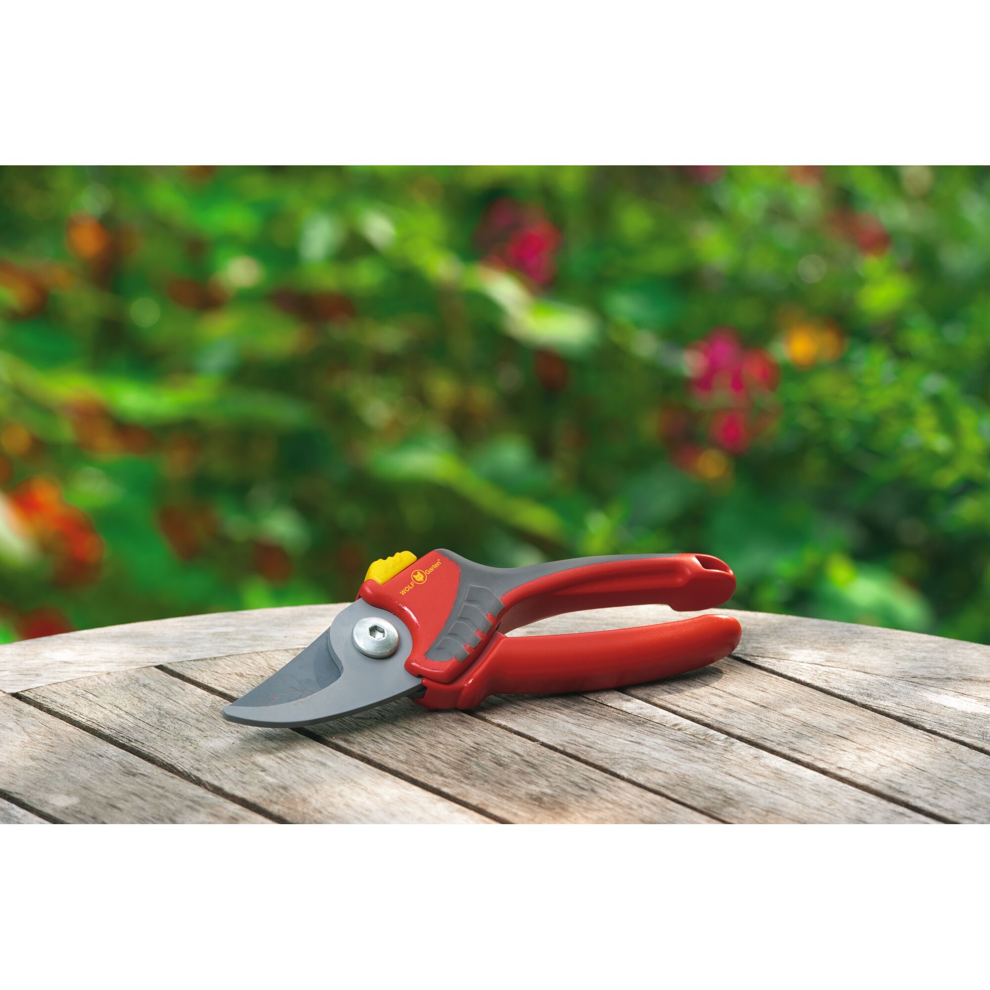 A pair of red and grey garden pruning shears from WOLFGarten is placed on a wooden outdoor table, with a blurred background of greenery and flowers.