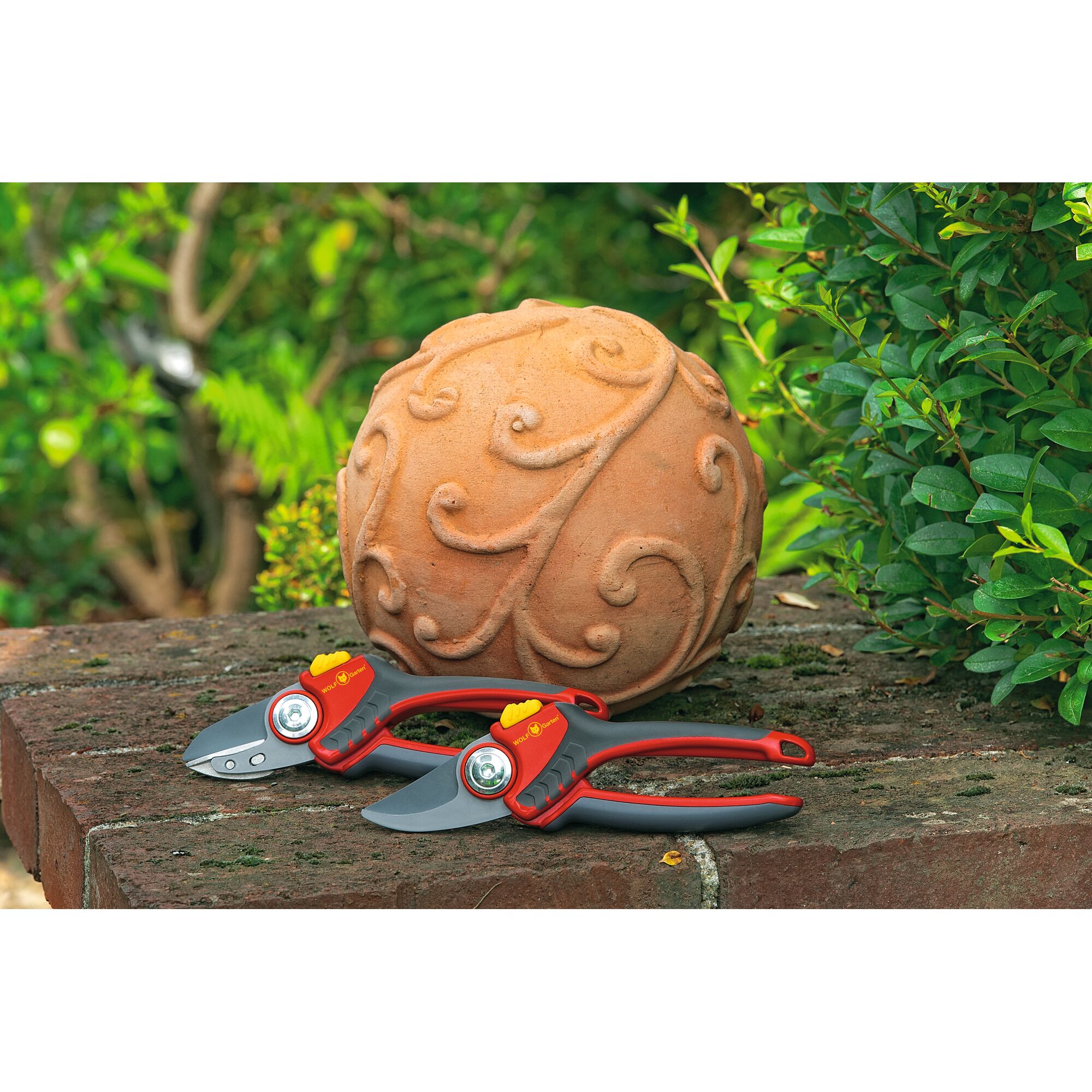 Two red and grey garden secateurs are placed on a brick ledge next to a decorative terracotta ball with a swirling pattern, surrounded by green foliage.