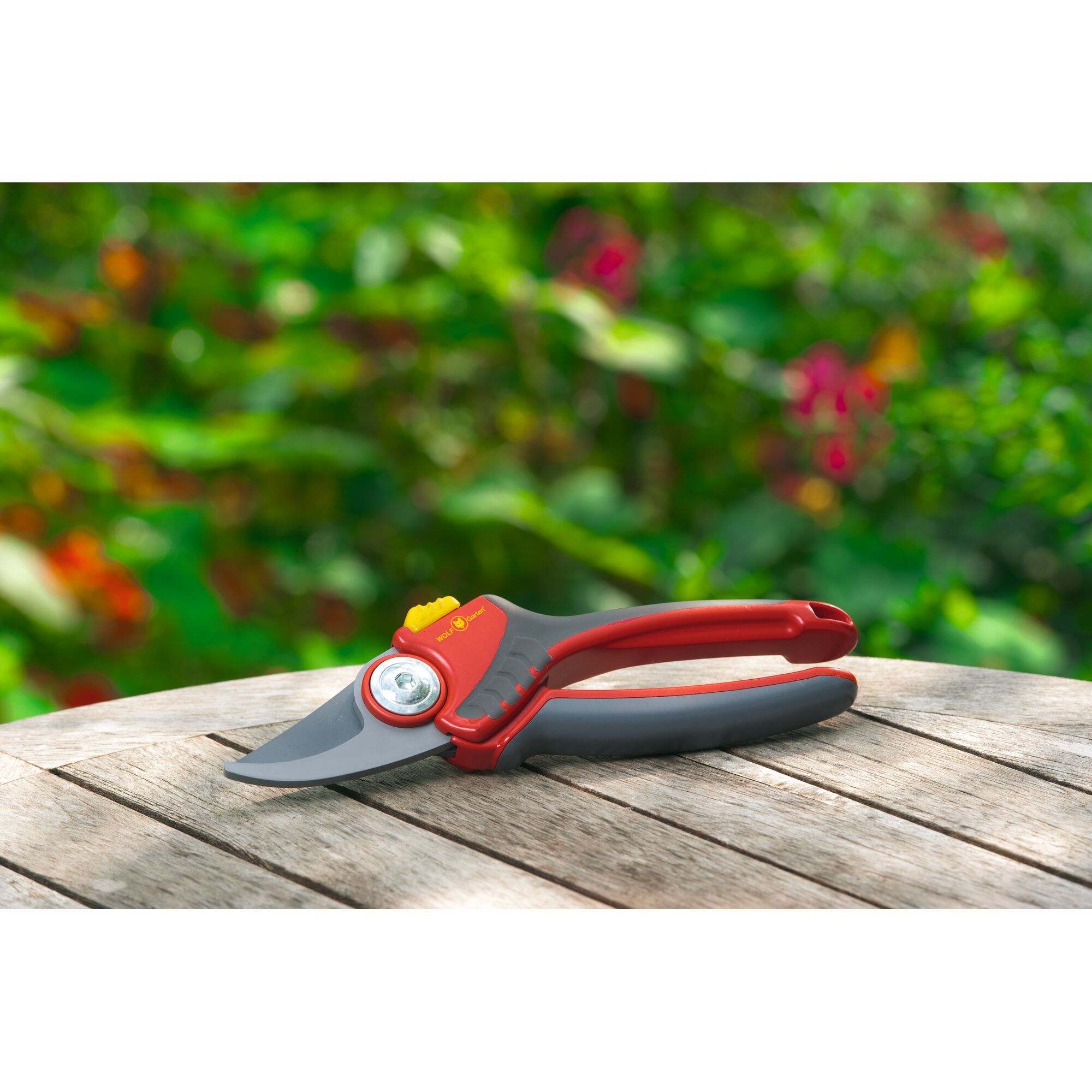 Red and gray garden pruning shears with a yellow button, placed on a wooden table outdoors with a blurred green foliage background.