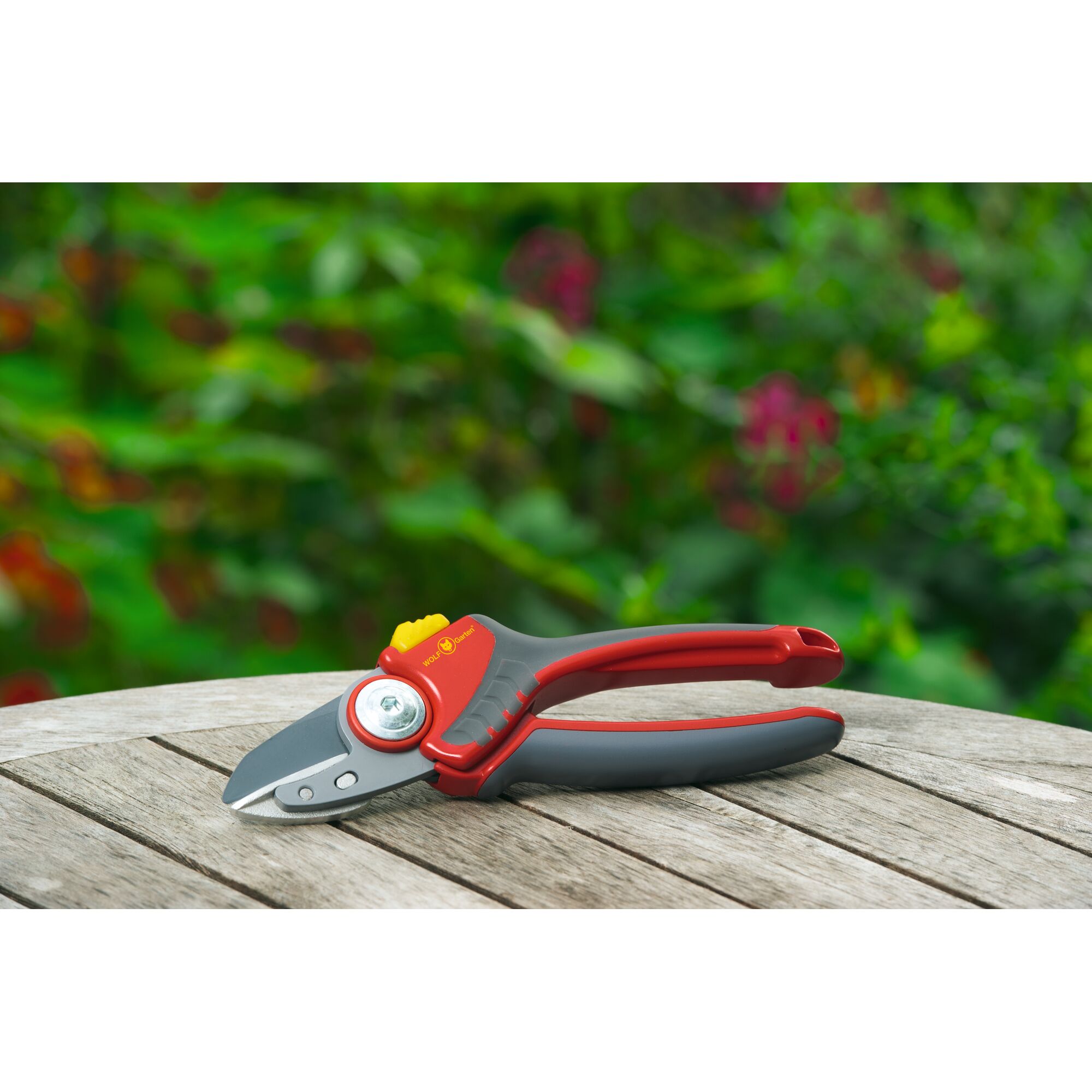 A pair of WOLF-Garten garden pruning shears with red and grey handles, resting on a wooden outdoor table, with a blurred green foliage background.